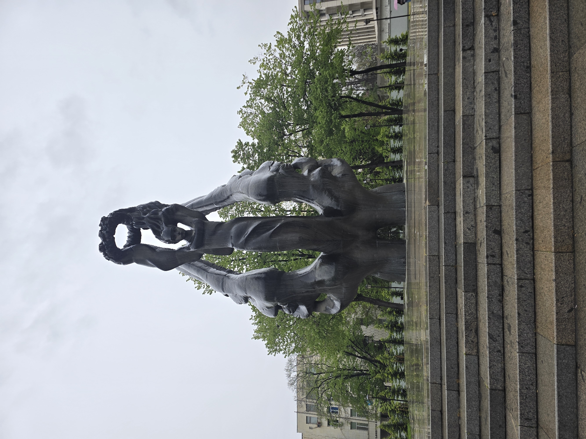 The image shows a sculptural monument with two figures holding a wreath in front of a building and steps in an urban environment. The figures are dark, possibly metallic, with a stylized, elongated form. The wreath appears to be made of leaves or flowers. Behind the monument are trees, a building with multiple windows, and a set of stone steps. The sky is overcast. The location is likely in an urban area of Unknown, Unknown, given the presence of the building and monument.