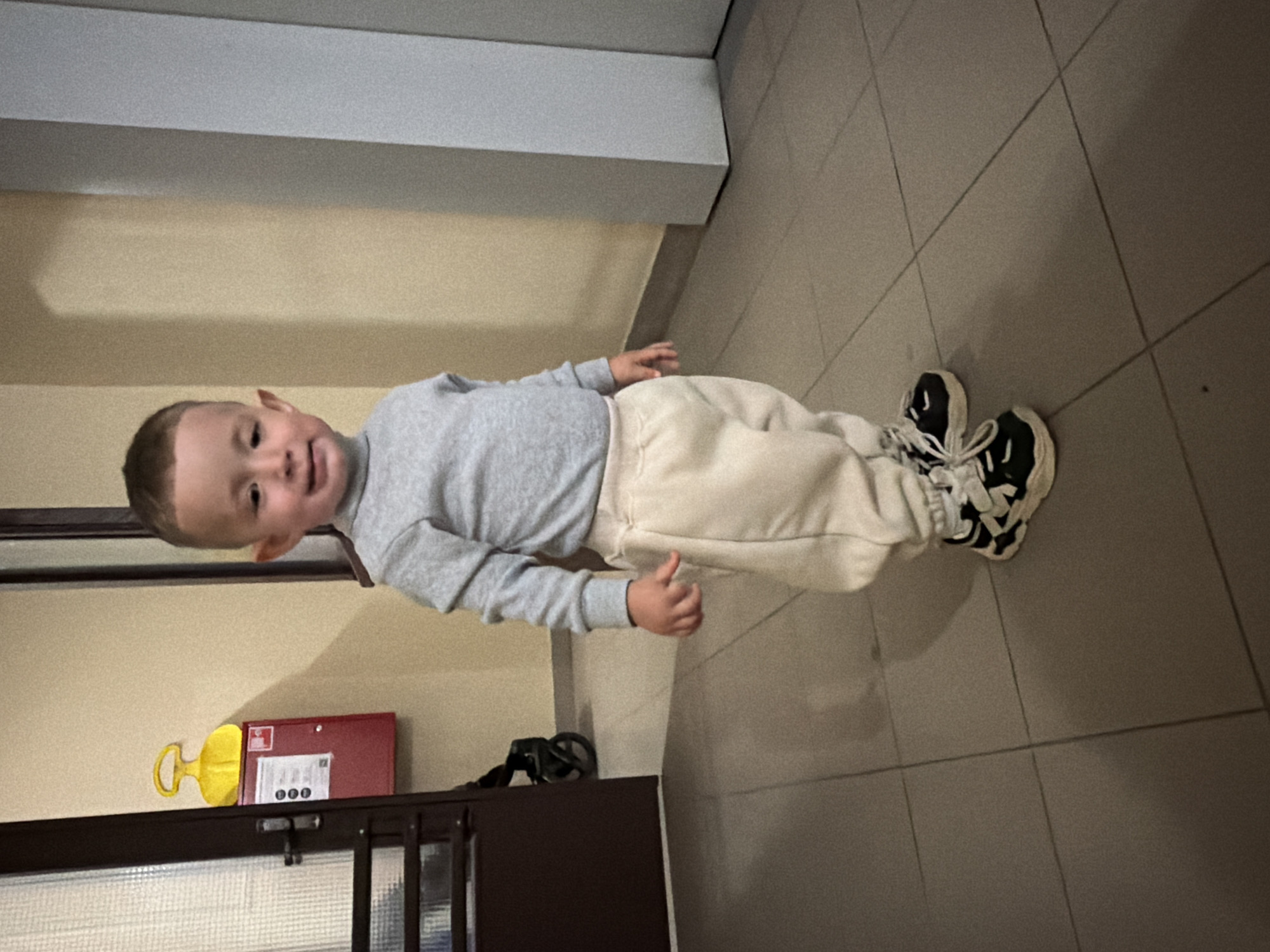 A young Caucasian boy, appearing to be around 2-3 years old, stands facing the camera. He has short, dark hair and is wearing a light grey turtleneck sweater and cream-colored sweatpants. His feet are adorned with black and white sneakers with prominent laces. The boy is positioned in what appears to be an indoor hallway or entryway, with tiled flooring. To the left of the boy, a dark brown door with a glass pane is visible, next to a red fire alarm box with a yellow handle. In the background, a wall with a slight yellowish hue is present, along with a glimpse of a white structural element on the right side of the frame. The lighting in the scene is subdued. The location is the city of Odintsovo, Russia.