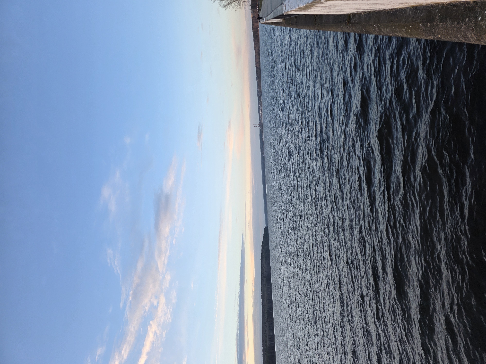 The image shows a waterscape in Chaikovsky, Russia. To the right, dark, choppy water fills approximately half of the frame, with the edge of a concrete pier or walkway visible at the very top. To the left, a bright blue sky with scattered clouds fills the other half of the frame. In the distance, across the water, there is a narrow strip of land or shoreline, possibly with trees or buildings visible along the horizon. The overall impression is one of a calm, peaceful environment by a body of water.