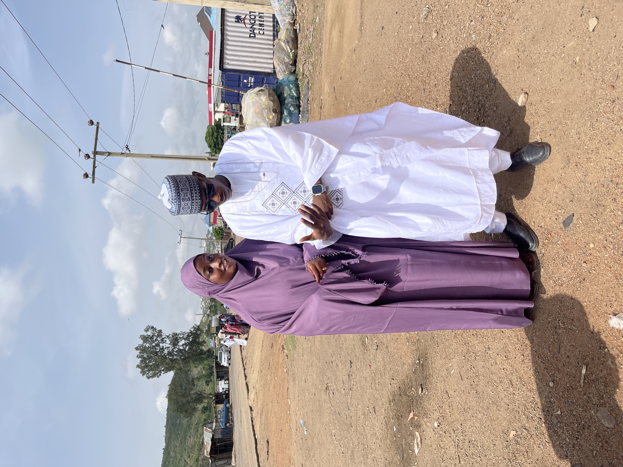 A man and woman are standing next to each other on a dirt road in Naraguta Hausawa, Nigeria. The man is wearing a white traditional outfit and a patterned hat, along with sunglasses. The woman is wearing a purple hijab. The background includes a road, a small hillside, and a few structures, along with utility poles and wires. In the distance is a container with "DANGOT CEMENT" printed on it.