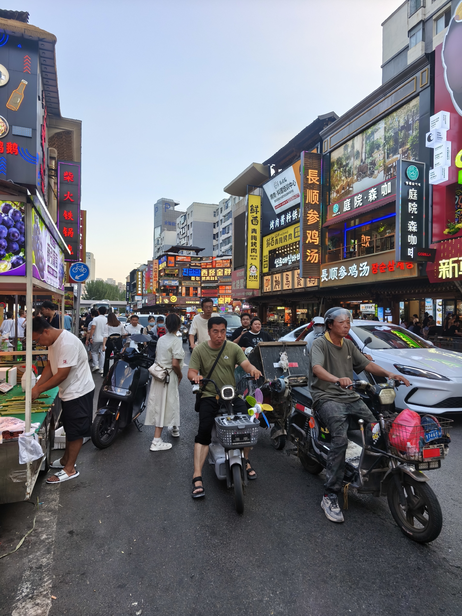 The image depicts a busy street scene in Heping District, China. Multiple individuals are present, primarily on foot or riding electric scooters and motorcycles. The street is lined with buildings adorned with numerous neon signs, many of which are in Chinese characters, indicating commercial establishments such as restaurants and shops. A white car is visible on the right side of the frame, suggesting vehicular traffic. People are engaged in various activities, including walking, riding, and seemingly interacting at a market stall on the left side of the image. The overall atmosphere suggests a vibrant urban environment during daylight hours.