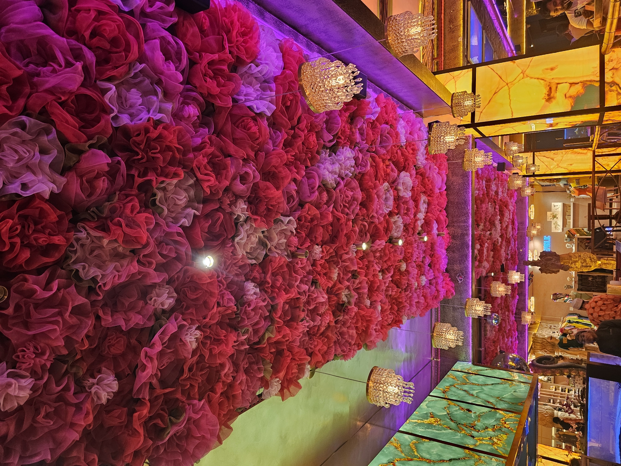 A vertically oriented, high-angle photograph showcases a decorative interior in Armação Dos Búzios, Brazil. Dominating the left side is a textured wall covered densely with fabric flowers in shades of pink, red, and light purple. Small, ornate chandeliers are suspended from the ceiling along the floral wall's edge.
To the right of the floral wall, the photograph captures a segment of the interior with a brightly lit, yellow-colored ceiling featuring a decorative pattern. Multiple chandeliers continue to be suspended from this ceiling as well.
A portion of the interior, including shelves stocked with items that may be merchandise, and people, can be seen in the background. In the foreground, there is a reflective, patterned surface in shades of green and gold.