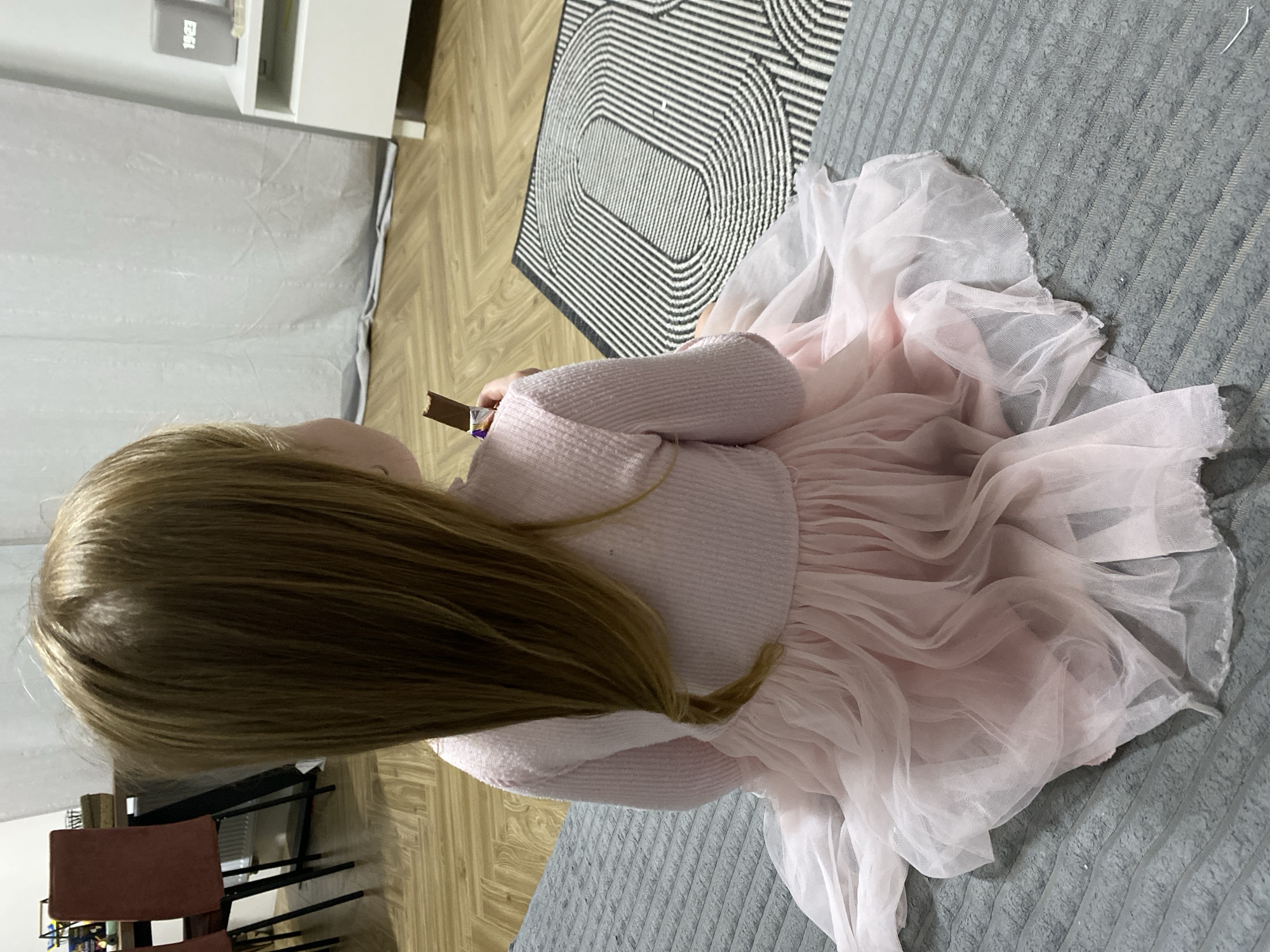 A young girl with long blonde hair sits on a grey patterned bed. She is wearing a light pink sweater and a layered pink tulle dress. The girl is holding a chocolate bar in her hands. In the background, there is a black and white rug on a wooden floor, as well as a white cabinet. The location cannot be determined from the image.