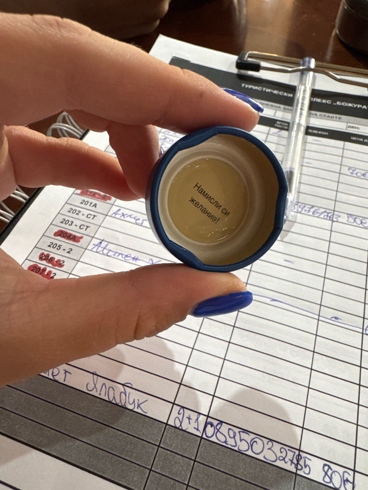 The image shows a close-up of a person's hands holding an open bottle cap. The inside of the cap has a yellow surface with text written in Cyrillic. The text reads "Намисли си желание!" which translates to "Make a wish!". The background is a grid-like paper, possibly a form or ledger, with some handwritten entries. There are also some printed headings in Cyrillic, including "ТУРИСТИЧЕСКИ" (Tourist) and "ЛЕКС „БОЖУРА" (Lex "Bozhura"). A pen is visible in the upper right quadrant of the image. Based on the printed text referencing "Bozhura", which is a hotel and restaurant complex located in Sredets, Bulgaria, it is highly probable that the scene is in or related to Sredets, Bulgaria. The overall impression is of a casual moment captured, possibly a playful interaction or a personal thought.