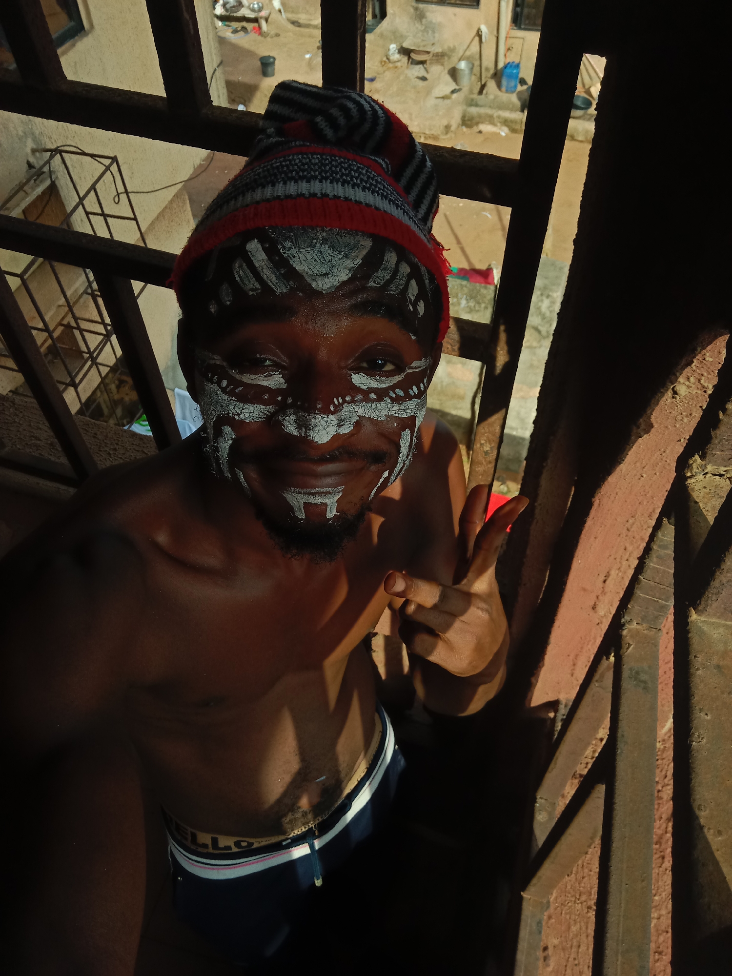 A medium-shot, high-angle photograph taken outdoors in Nkpor, Nigeria, shows a dark-skinned, shirtless man wearing a striped knit hat and underwear, standing on a rusted metal staircase. The man has white face paint on his forehead, around his eyes, and on his chin and upper lip. He is making a hand gesture with his right hand. The background shows a courtyard with various objects scattered around. The lighting in the photograph is uneven, creating strong contrasts between light and shadow.
