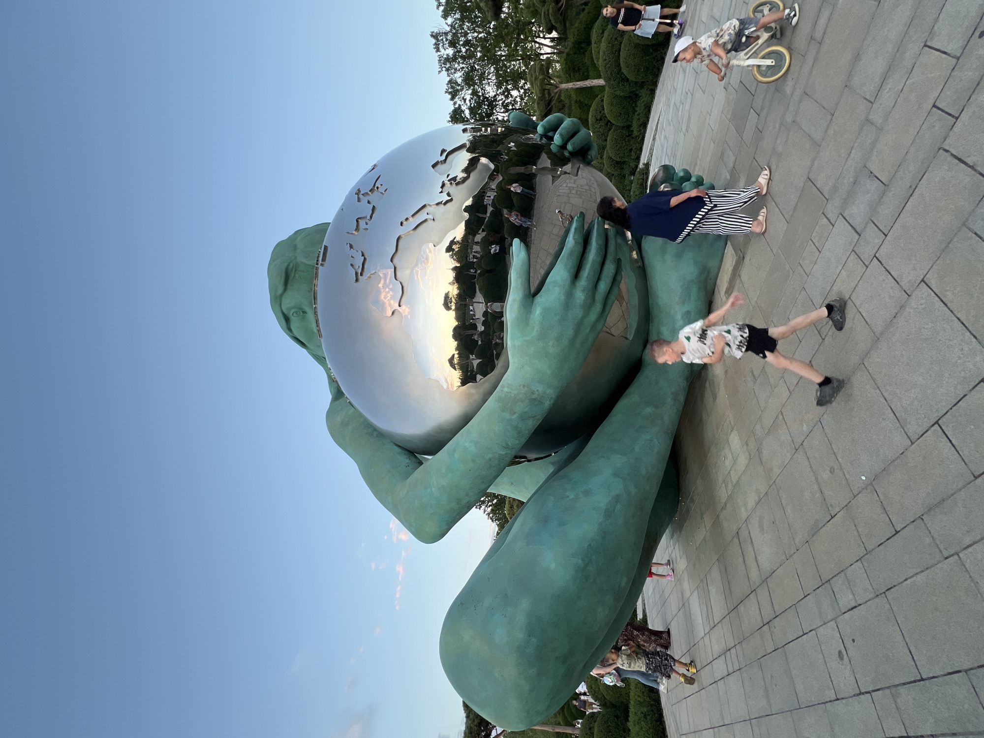 This media file captures a large, green-patinated bronze sculpture of a figure holding a mirrored globe. The globe reflects the surrounding environment, including trees and a cloudy sky with sunset hues. Several people are present around the sculpture. In the background, a young boy rides a bicycle on a paved walkway. Another child, wearing a patterned shirt and shorts, is captured in motion on the walkway, appearing to run or jump. A person wearing a dark top and striped pants stands near the sculpture. The scene appears to be outdoors in a public space, likely a park or plaza, given the sculpted hedges and paved ground. The lighting suggests it is late afternoon or early evening, as indicated by the warm tones in the sky's reflection. The overall mood seems relaxed and casual, with people enjoying the outdoor space. The location is identified as Krasnodar, Russia.