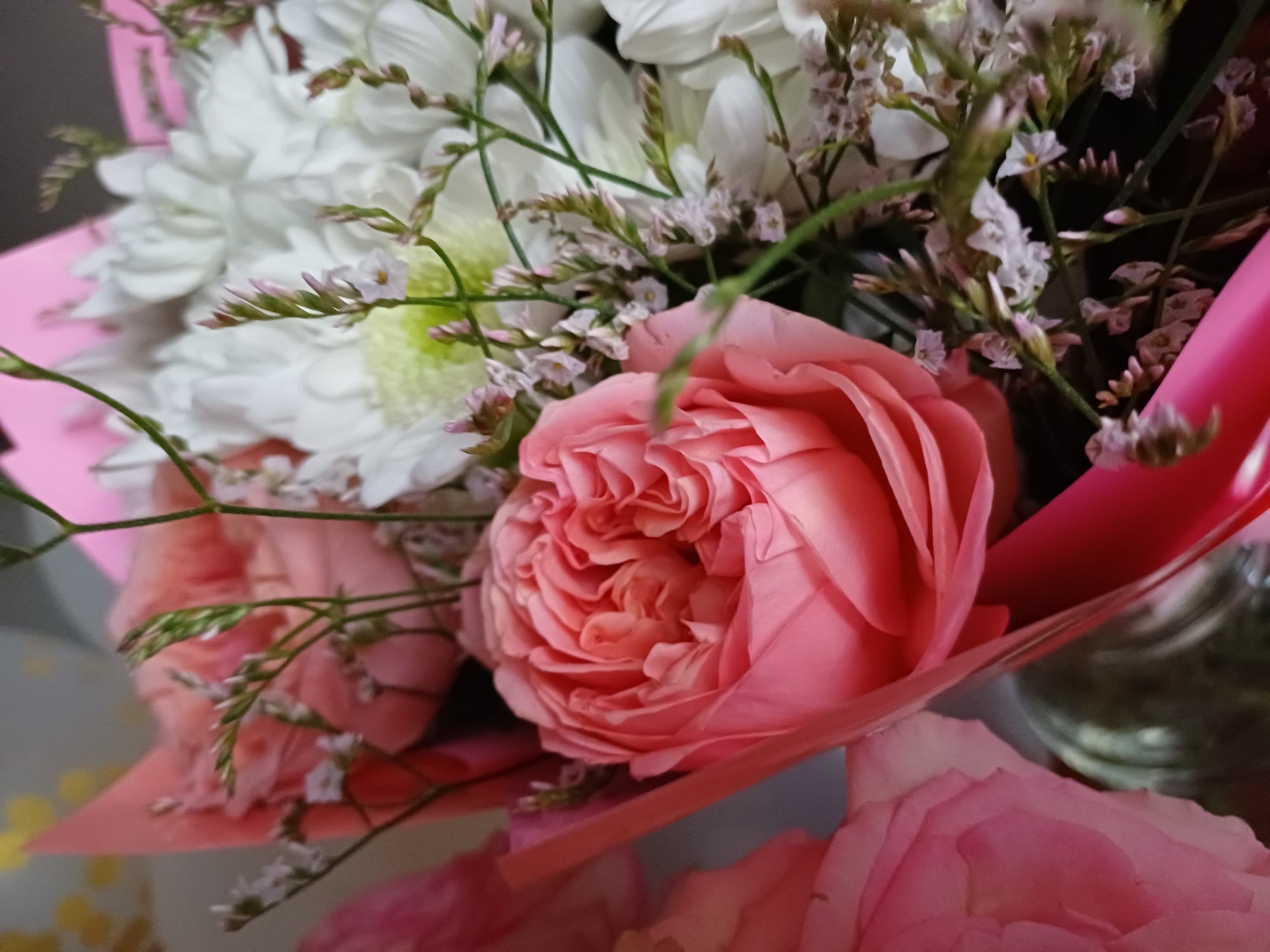 This is a close-up, eye-level shot of a bouquet of flowers. The primary subjects are a large, fully bloomed pink rose in the foreground, its petals tightly swirled and layered, and several white, chrysanthemum-like flowers in the background. Delicate, small, pale pink and white filler flowers and thin green stems are interspersed throughout the bouquet. The flowers are arranged with a vibrant pink wrapping that forms a cone-like shape around them. The lighting is soft and diffused, suggesting an indoor setting, possibly a room with natural light. The overall mood is delicate and romantic. There is no visible text in the image.