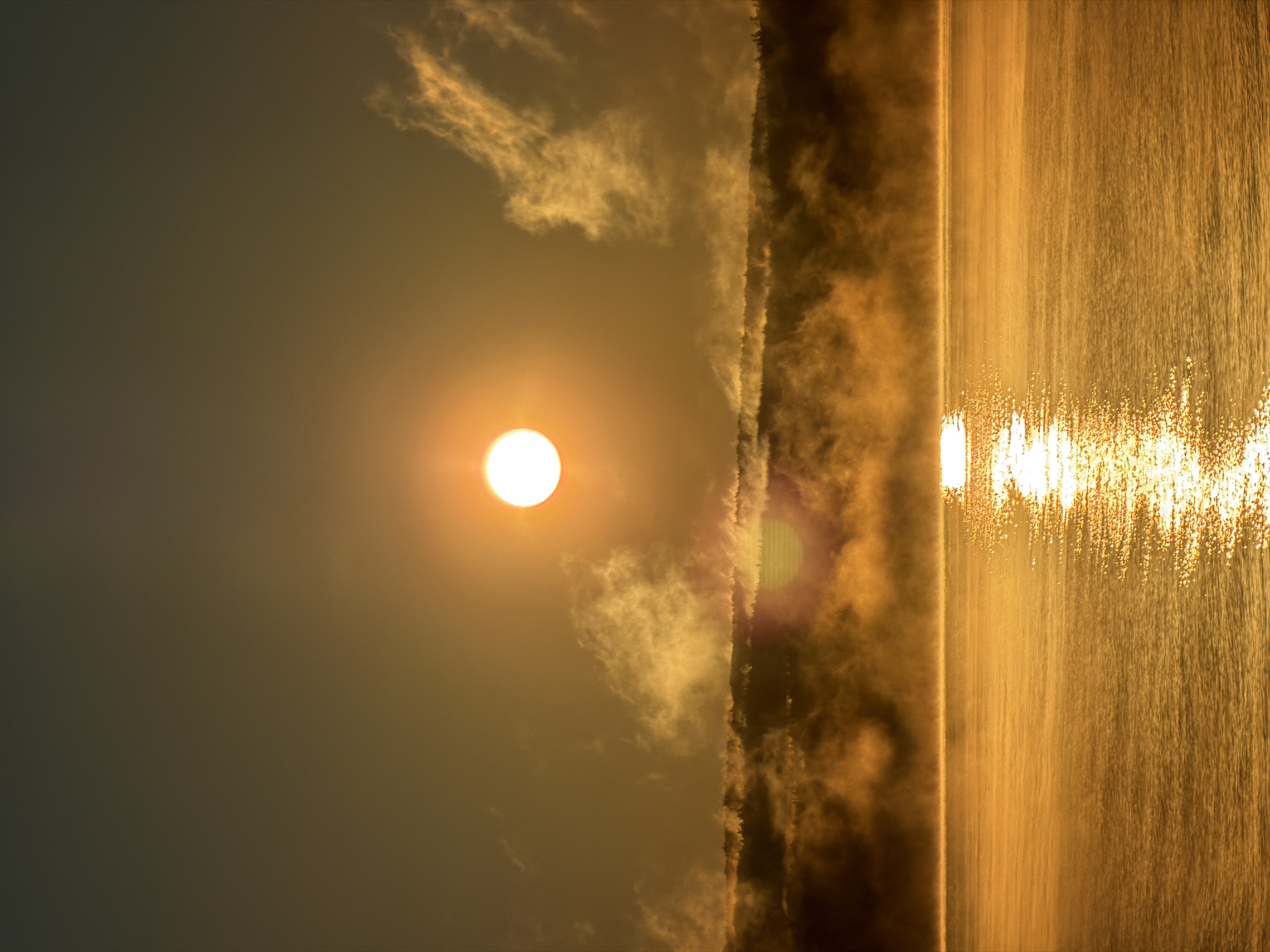 The image depicts a sunrise over a body of water, likely a reservoir or river, as seen from Chaikovsky, Russia. The sun is a bright, luminous orb positioned high in the sky, casting a warm, golden light across the scene. Wisps of fog or mist rise from the surface of the water, partially obscuring a treeline in the background. The water itself reflects the sunlight, creating a shimmering, textured surface. The sky is a gradient of warm tones, transitioning from a deep orange near the horizon to a muted gray at the top. There are no people or specific man-made structures visible in the image, emphasizing the natural landscape. The overall impression is one of serene, early morning tranquility.