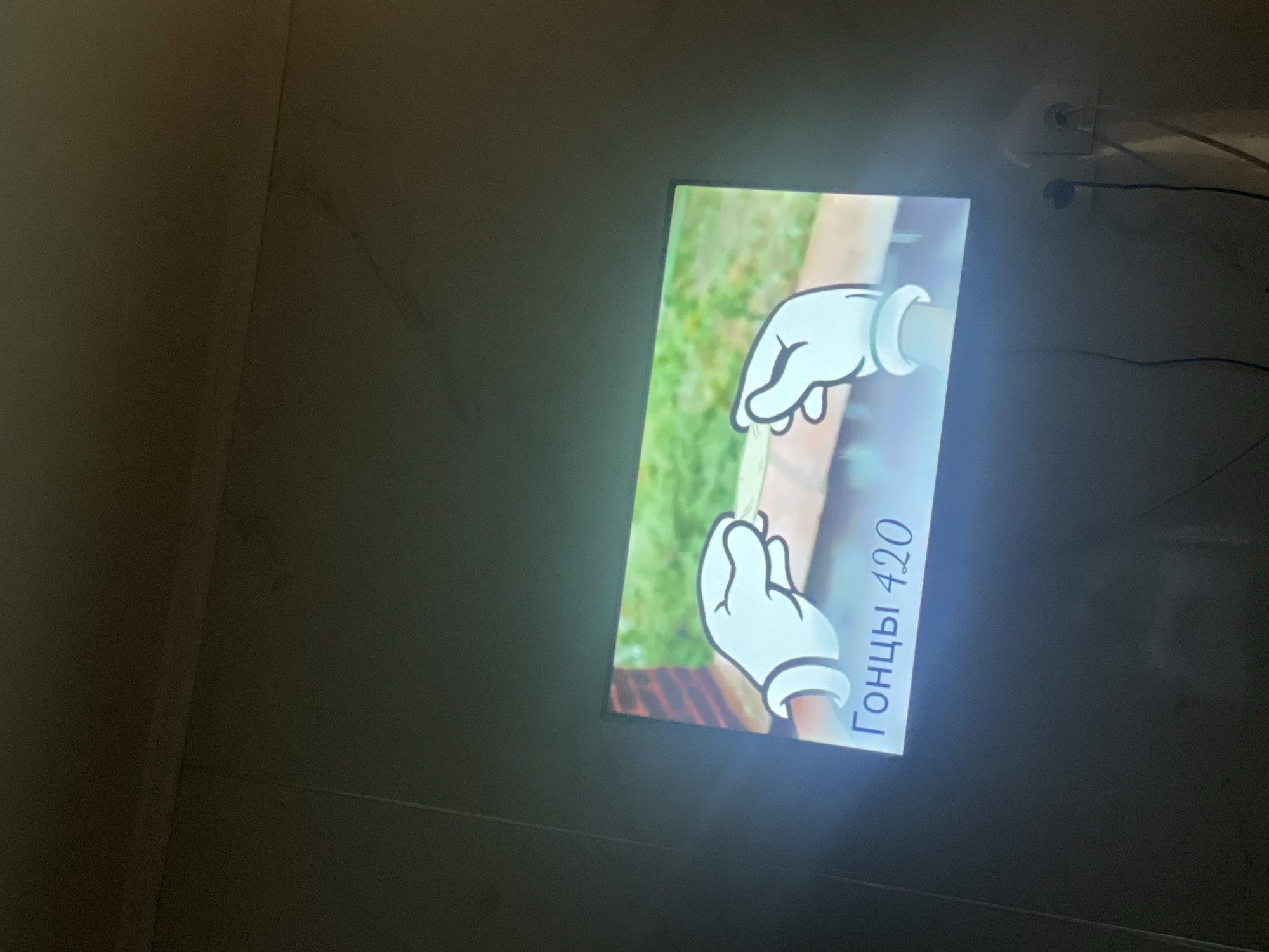 An illuminated television screen displays an animated scene featuring two cartoon hands wearing white gloves, positioned as if holding something delicate. Below the hands, in blue Cyrillic text, are the words "Гонцы 420". The television is mounted on a wall, with electrical cords and outlets visible in the lower right portion of the image. The surrounding environment appears to be a dimly lit room. The city of Kojomkul, Kyrgyzstan, is mentioned in the context of this scene.