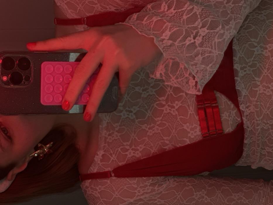 A woman's perspective is captured in a selfie. The image is lit with red light. Her left hand holds a smartphone with a glittery grey case and a pink, circular-patterned grip. She's wearing a white lace garment over a red piece of clothing. Her hair is reddish-brown and falls over her left shoulder, displaying a gold and red earring. The focus is on the hand, phone, and the lace material.