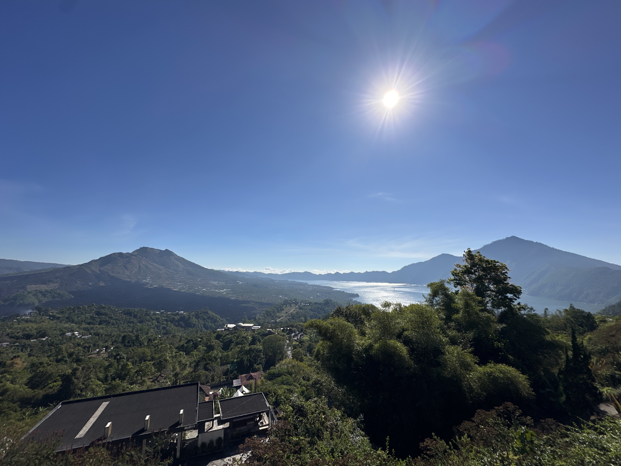 The image is a panoramic view of the Kintamani region in Bali, Indonesia, on a clear, sunny day. The foreground features the dark, sloped roof of a modern building, partially obscured by lush green foliage. Beyond the building, a dense, verdant landscape of trees and scattered houses stretches towards the middle ground.
Dominating the mid-ground and background is a vast, tranquil lake, likely Lake Batur, reflecting the bright sunlight and the hazy outlines of distant mountains. On the left side of the lake, a large, dark volcanic mountain rises prominently, its slopes appearing barren in some areas and covered in vegetation in others. To the right of the lake, more rounded, green-covered mountains form a picturesque backdrop.
The sky is a brilliant, cloudless blue, with the sun positioned high in the upper right quadrant, casting a bright, starburst-like glare. The overall impression is one of serene natural beauty, showcasing the volcanic landscape and the expansive lake characteristic of Kintamani. There are no people visible in the image, and the scene is static, depicting a landscape at a specific moment in time. The environment is natural and mountainous, with evidence of human habitation in the form of buildings and scattered structures.
