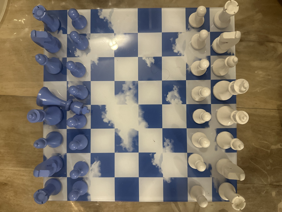 A chessboard, its squares alternating blue and white with a sky and clouds pattern, is set up for a game. On one side, a full set of periwinkle blue chess pieces is positioned, while on the opposite side, a full set of white pieces mirrors the arrangement. The board and pieces are situated on a surface, slightly angled from an overhead view.