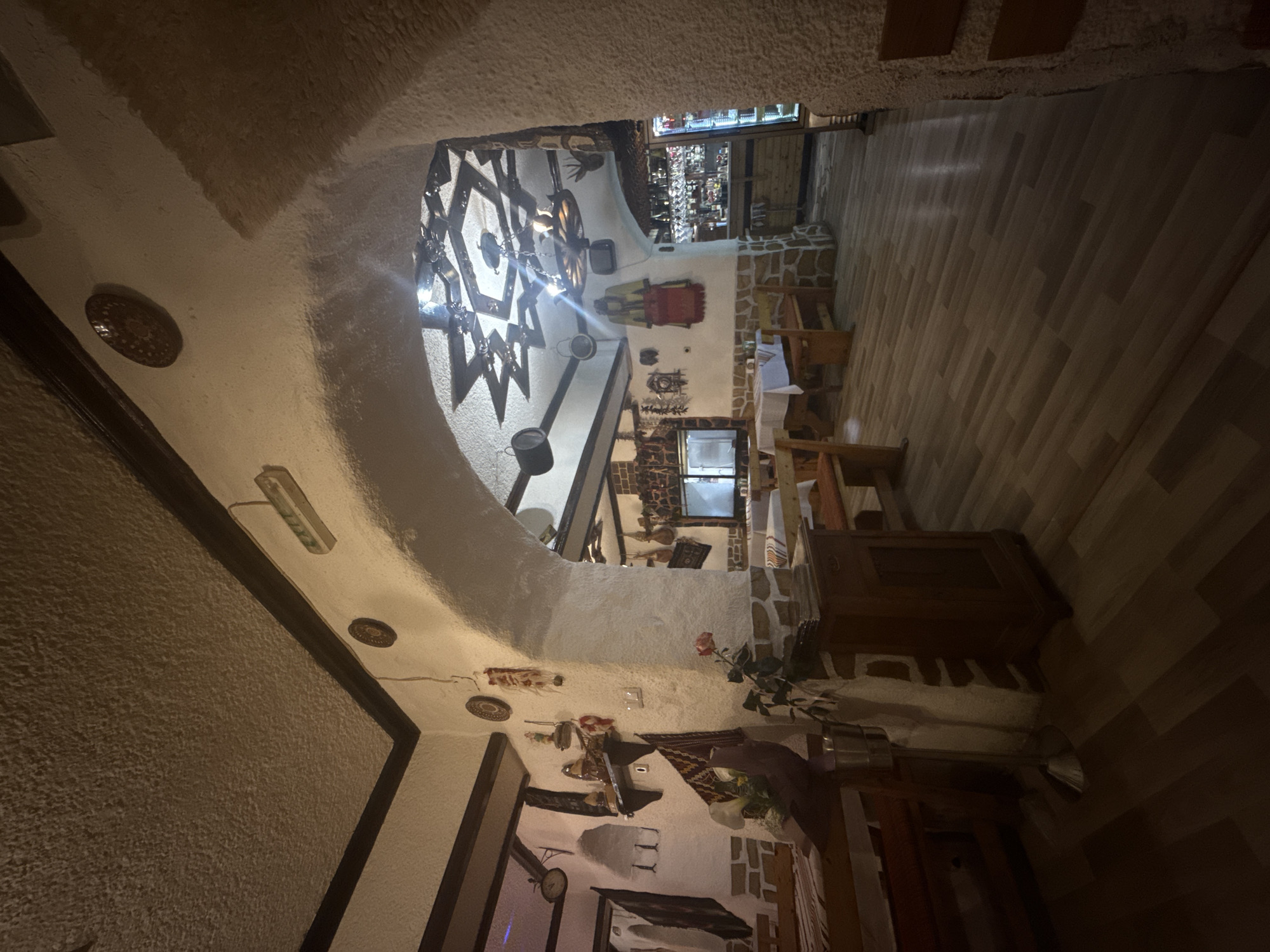 The image depicts the interior of a restaurant in Varna, Bulgaria. The scene is shot from a low angle, looking upwards and into the restaurant. The ceiling is textured and arched, with decorative plates and light fixtures. A large, ornate chandelier hangs in the center. The walls are whitewashed and adorned with various decorative items, including rugs, pottery, and mounted objects. Tables and chairs are arranged in a dining area, with a bar visible in the background. The floor is made of wooden planks. There are no people visible in the frame.