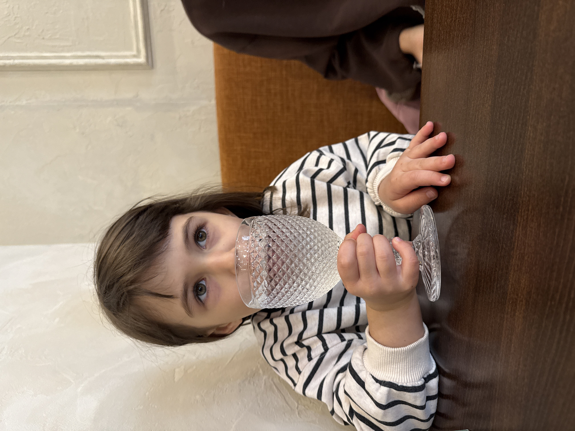A young girl in a black and white striped shirt holds a glass with a textured surface to her mouth. The scene is indoors. The girl is seated at a dark brown wooden table. Part of an orange chair is visible. Another person wearing a brown sweater is partially visible in the background. A textured, light-colored wall is behind the girl, with a framed section. The photo was taken in Krasnoyarsk, Russia.
