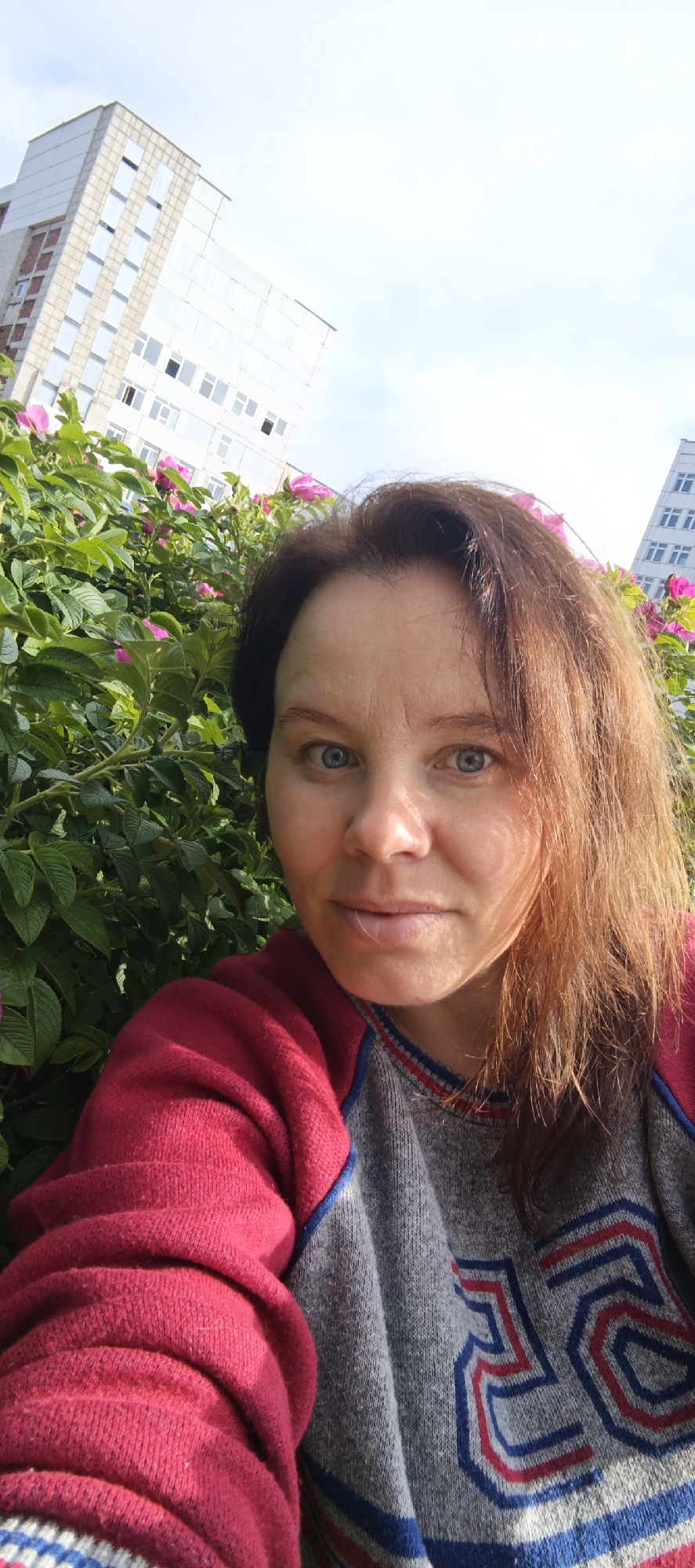 A selfie taken in Chaikovsky, Russia, shows a woman with shoulder-length auburn hair and blue eyes. She is wearing a red sweater over a grey sweater with blue and red stripes and the number "25" emblazoned on it. She is positioned in front of a lush green bush with pink flowers. In the background, modern buildings with numerous windows are visible against a clear blue sky, suggesting an urban setting. The lighting indicates it is daytime, likely with sunlight casting shadows on the woman's face.
