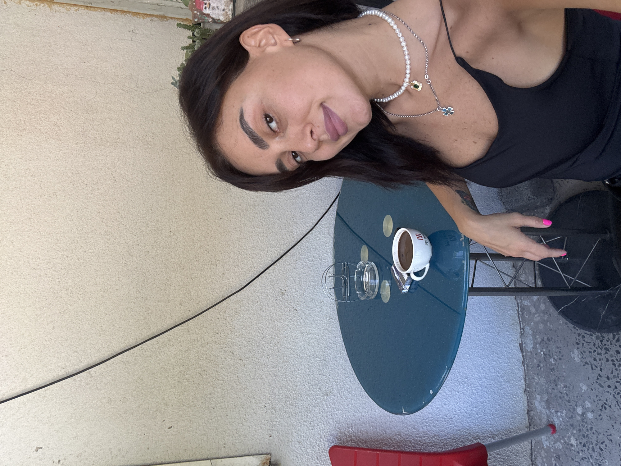 A woman with dark hair, wearing a black tank top and two necklaces, is sitting at a small blue table with a cup of coffee and a pack of cigarettes. She is looking at the camera. The table is outdoors, with a textured wall in the background. A red chair is visible to the left of the table. The scene appears to be in Tbilisi, Georgia, given the context provided.