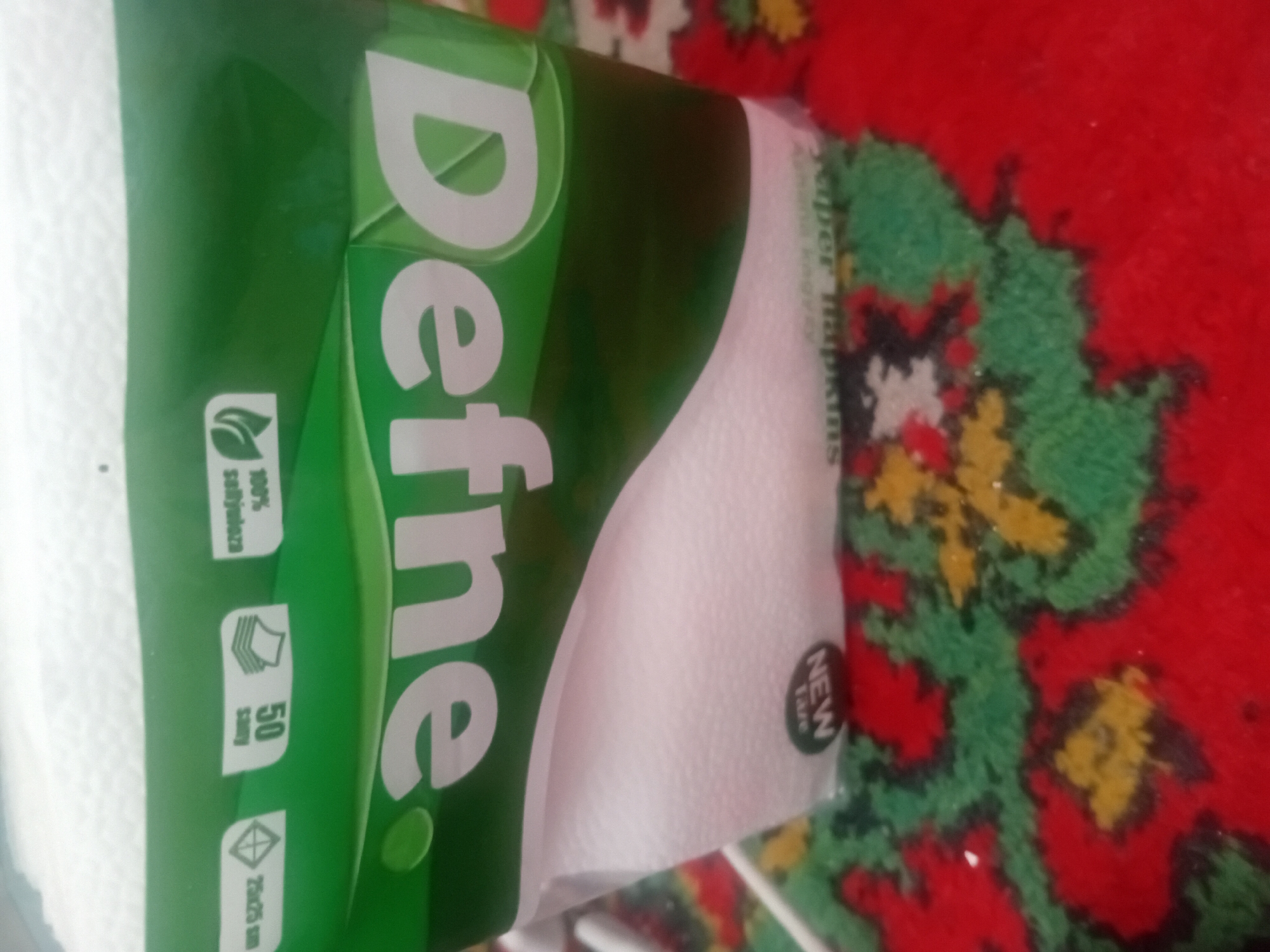 The image shows a close-up of a pack of "Define" paper napkins. The packaging is primarily green and white. On the green band, there are several icons and text, including "25x25 sm", "50", and "100% softyloza", along with a leaf icon. The word "Define" is prominently displayed in large white letters across the front of the pack. At the bottom of the pack, there is a circular "NEW Tare" label. The pack is placed on a red, green, and yellow patterned rug. The background is blurred but appears to be an indoor setting. There are no people visible in the image. The image is a product shot, focusing on the packaging of the paper napkins. There is no information within the image that would indicate a specific location in the city of Ashgabat, Turkmenistan.