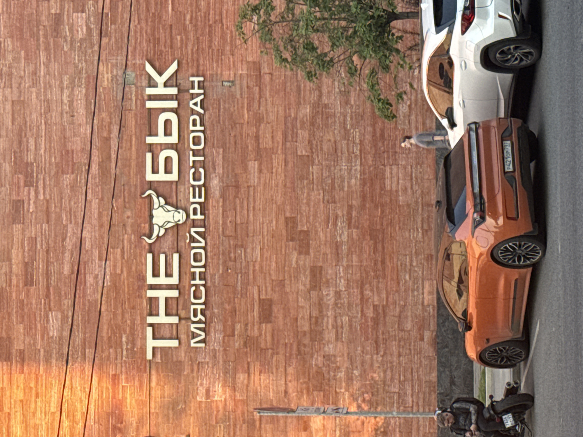 The image depicts the exterior of "The Bull" restaurant in Moscow, Russia. The restaurant's sign, featuring the name in Cyrillic and an image of a bull's head, is visible on the brick façade of the building. Two cars, an orange SUV with the license plate P421OP 777 and a white SUV, are parked next to the building. A man is standing near the white car. A motorcyclist sits on his motorbike at the end of the sidewalk.