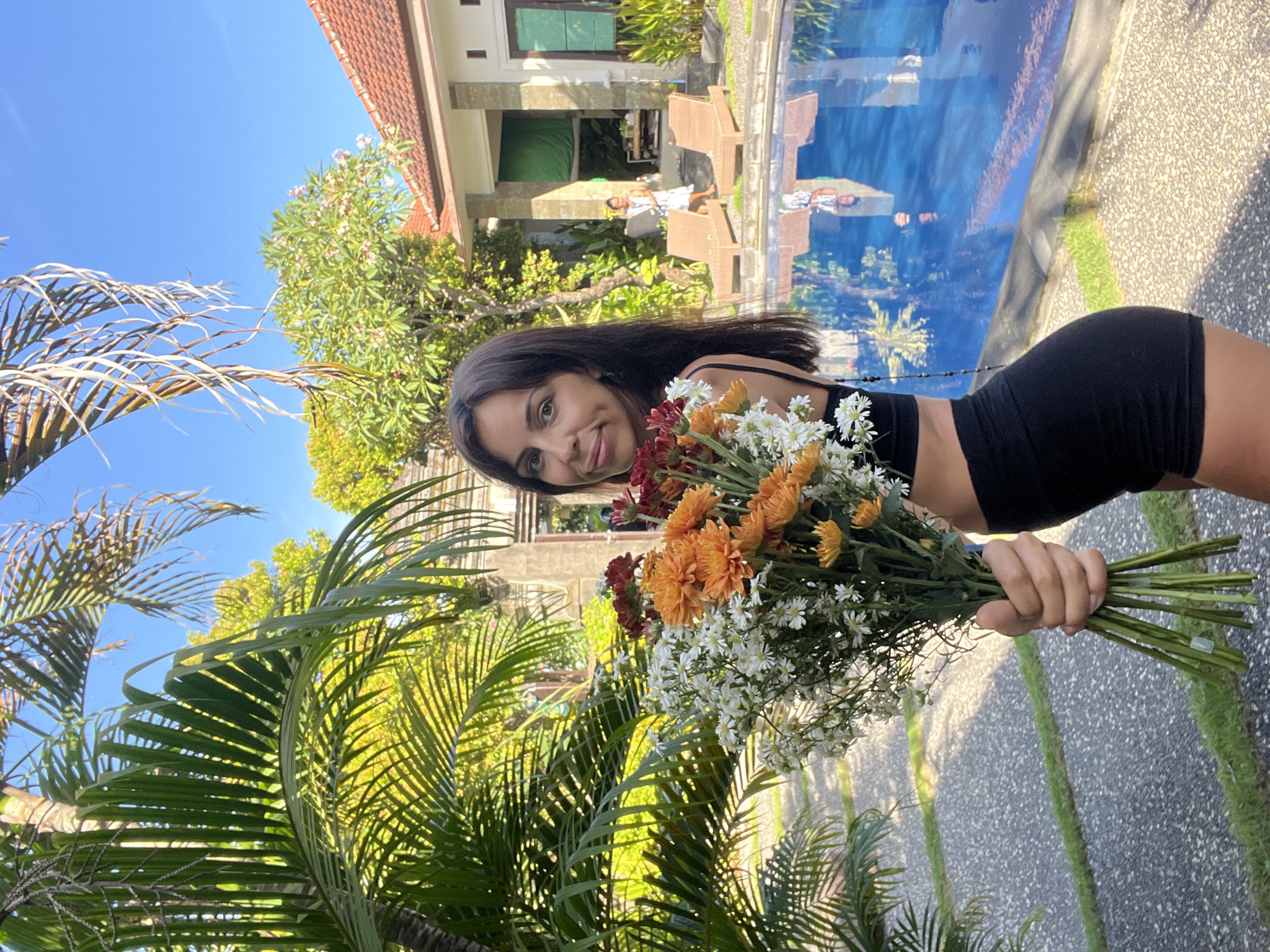 A young woman is captured in a vibrant outdoor scene, holding a large bouquet of fresh flowers. She has long dark hair, dark eyes, and a pleasant smile on her face. She is wearing a black sleeveless top and black shorts. The bouquet in her left hand consists primarily of white daisies, bright orange chrysanthemums, and some reddish-brown blossoms, with prominent green stems.
The setting appears to be a tropical resort or villa in Kuta Selatan, Indonesia, indicated by the lush greenery and architecture. Behind her, dense green foliage and palm trees create a natural backdrop. To her right, a clear blue swimming pool reflects the sky and parts of the nearby buildings, with lounge chairs visible on the deck. Another individual in light clothing can be seen standing near the pool in the distance. To her left, bright blue sky is visible above the palm fronds, some of which appear dry and light-colored, while others are vibrantly green. The ground beneath her is a light-colored, textured paved surface with narrow green strips. The scene is bathed in bright daylight, suggesting a sunny day. No visible text is present in the image.