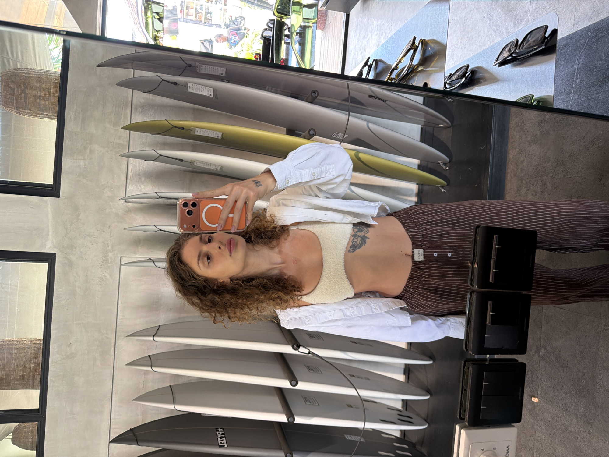 The image depicts a person taking a selfie in a mirror reflecting a surfboard shop. The person has curly brown hair and is wearing a white shirt and striped pants. The reflection shows multiple surfboards displayed on a wall. The shelf above the surfboards holds sunglasses. The environment suggests an indoor retail setting. The surf shop is located in North Kuta, Indonesia.