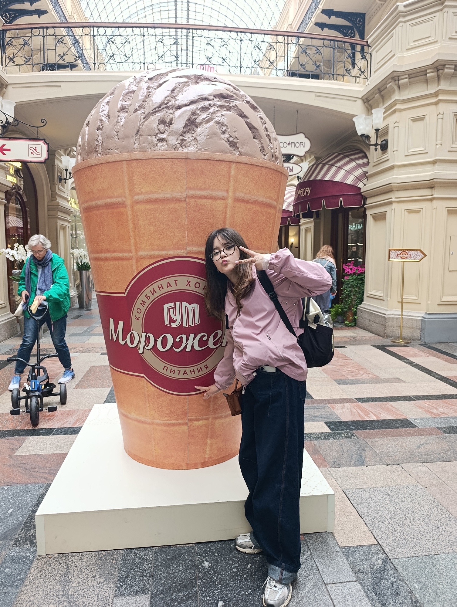 A young woman with dark hair and glasses is posing for a photo in front of a giant ice cream cone sculpture. She is wearing a pink jacket, dark jeans, and sneakers. She is making a peace sign with her left hand. To her left, an older person with gray hair is riding a three-wheeled bicycle and cleaning it. The scene is set indoors, likely a shopping mall or market, with arched ceilings and decorative architecture. The floor is tiled with a geometric pattern of pink, gray, and black. In the background, there are shops and signage, including a directional arrow sign with an image of what appears to be a flower.
