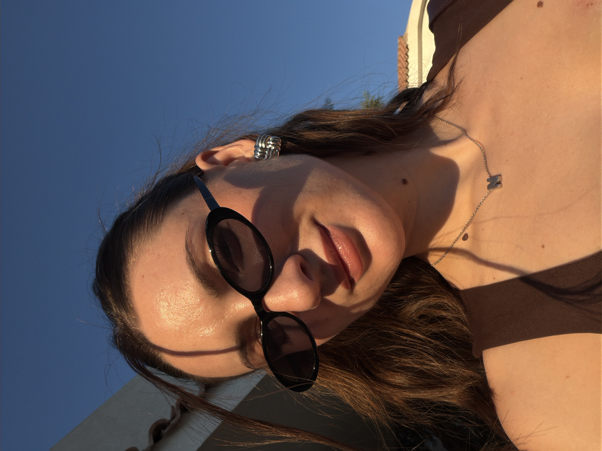 A young woman with dark brown hair is taking a selfie outdoors against a clear blue sky. She is wearing large, oval-shaped, black sunglasses and large, silver, twisted hoop earrings. A thin silver necklace with a single letter 'N' pendant rests on her chest. She is wearing a dark brown tank top. The lighting suggests it is late afternoon, with warm sunlight casting shadows. In the background, the edge of a building with a tiled roof is visible. The environment is consistent with a sunny, outdoor location in Sharm El Sheikh, Egypt.