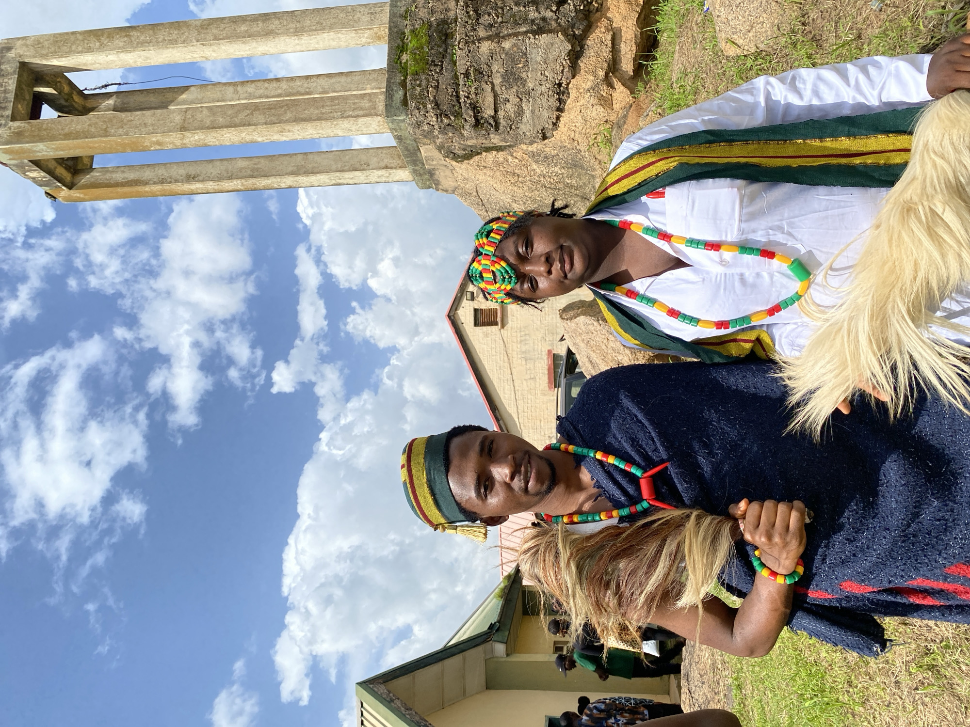 The image depicts two individuals adorned in traditional attire, likely participating in a cultural event or ceremony in Wang, Nigeria. The subjects are a man and a woman, both wearing colorful beaded necklaces and bracelets in red, yellow, and green. The woman is wearing a white shirt with a green and yellow sash draped across her shoulders. The man is wearing a dark blue garment over a white shirt and a cap with similar colorful accents.
The setting appears to be outdoors, with a backdrop of a rocky hill and a cloudy blue sky. In the foreground, lush green grass is visible. A concrete structure, possibly part of an ancient monument or building, stands to the left. The sunlight suggests it is daytime, and the clouds indicate it might be a partly cloudy day. The overall atmosphere seems festive and celebratory, with the subjects looking relaxed and engaged.
The image is oriented vertically. The people are positioned in the center, with the natural landscape and structures surrounding them. The bright colors of their clothing and accessories stand out against the more muted tones of the environment. The expressions on their faces suggest a sense of pride and enjoyment. No visible text is present in the image.