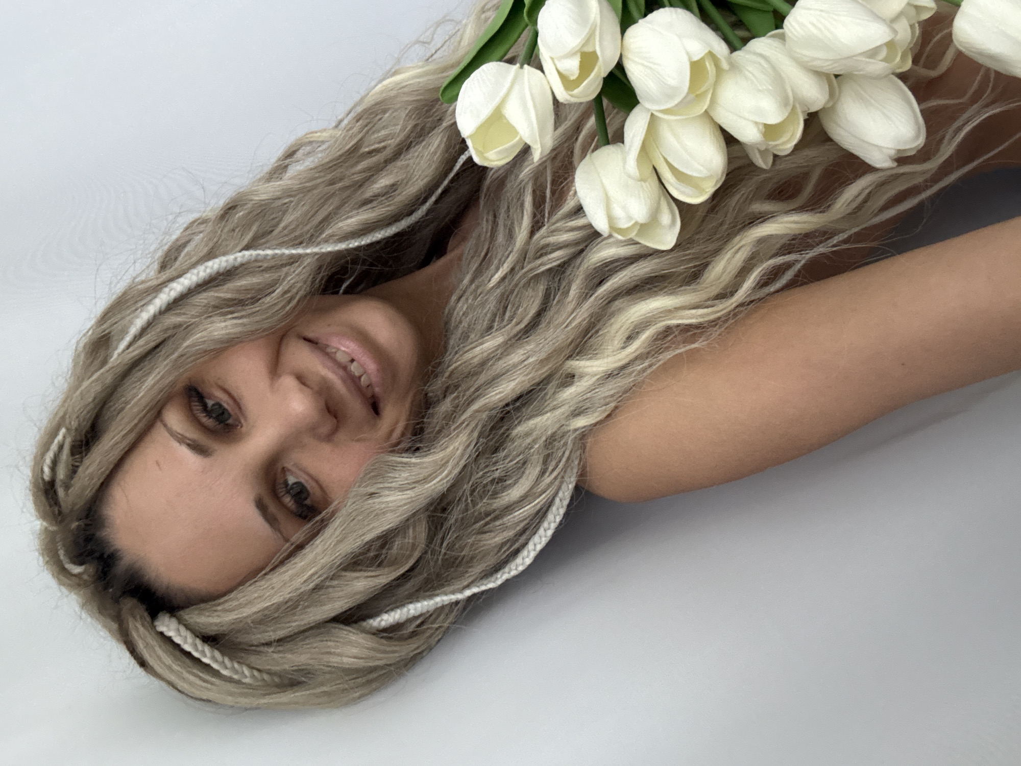 An overhead shot captures a smiling young woman with long, wavy, multi-toned blonde hair, possibly with light brown undertones, lying against a plain light grey background. Her head is positioned towards the left of the frame, angled slightly, and she looks directly up at the camera with a gentle, joyful smile. Her eyes are light-colored, and she has well-defined eyebrows. Several thin, white braided extensions are intricately woven throughout her hair, framing her face and blending with her long locks.
A fresh bouquet of white tulips with vibrant green stems is placed above her head, resting on her hair and extending towards the upper right corner of the image. The tulips are tightly closed, typical of their early bloom stage, and appear pristine. The woman's bare arm, with fair skin, is visible extending from behind her head towards the right side of the frame. The lighting is bright and even, creating soft shadows and highlighting the textures of her hair and the tulips. The overall mood is serene and cheerful, suggesting a connection to nature or a celebration. No text is visible in the image. The context for this media file is Rusanovo, Russia.