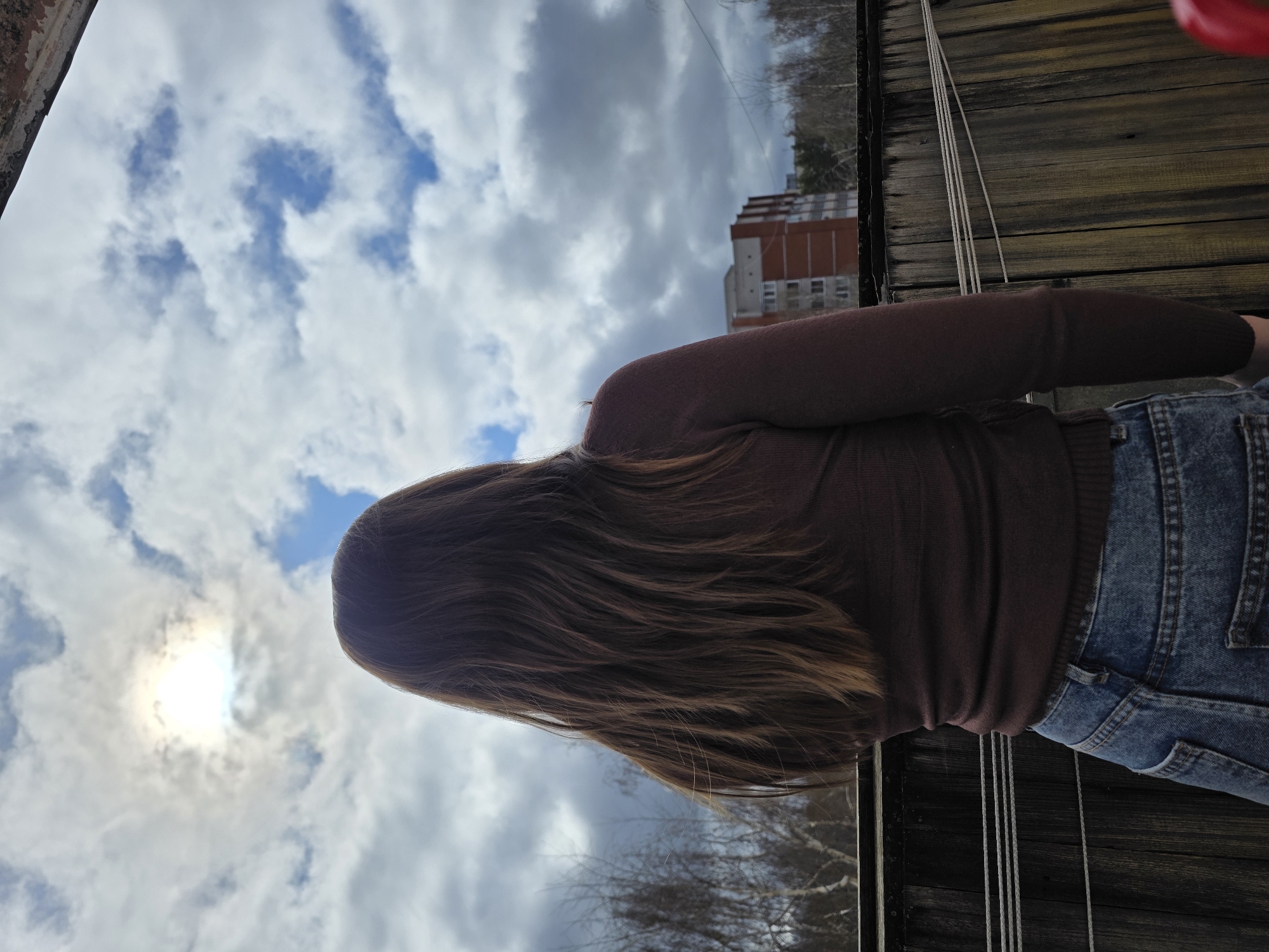 The image depicts a person with long, brown hair, wearing a brown sweater and blue jeans. The person appears to be standing on a wooden structure, possibly a balcony or deck. In the background, there is a cloudy sky with patches of blue and a building. The location is identified as Chaikovsky, Russia.