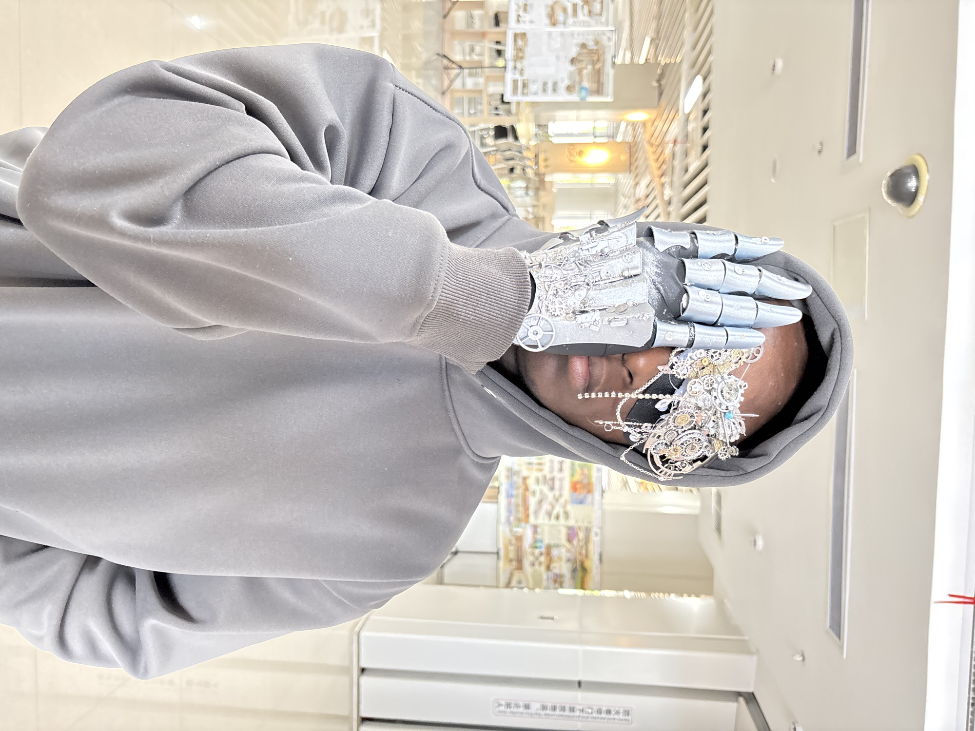 A person wearing a gray hoodie with a metallic hand covering their face is partially visible in this image. The person's hand is adorned with intricate metallic and gear-like attachments. They are also wearing a decorative metal headpiece that peeks out from underneath the hood. The background appears to be a store or a commercial space with shelves containing various items. There is a white wall with air conditioning vents and other room fixtures present as well. While the exact location cannot be verified, the image may contain writing in the Chinese language.