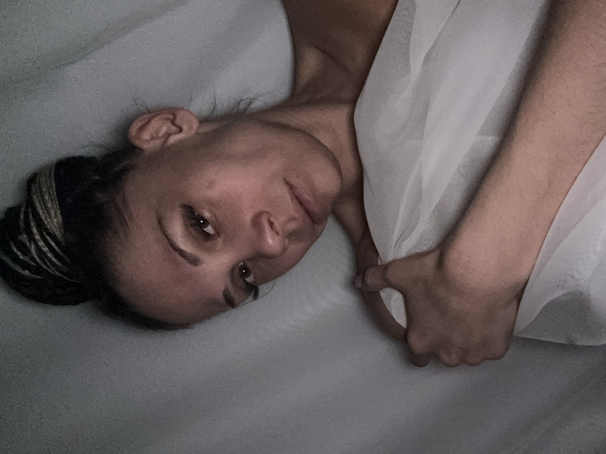 A woman with braided hair tied in a bun is lying down, partially covered with a white cloth. She has fair skin and is looking towards the camera. The background is a muted gray. There is no evidence of a city or location to confirm it is Rusanovo, Russia.