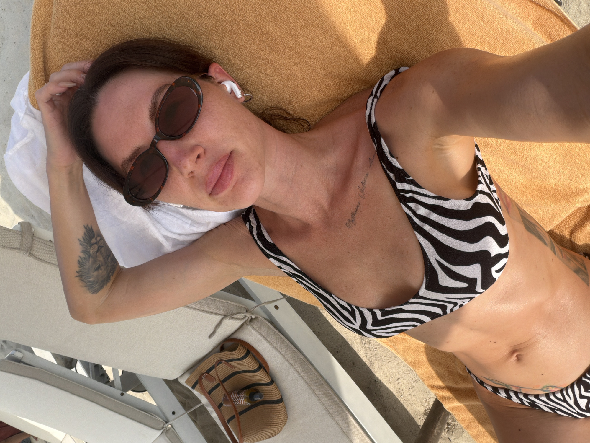 A woman with dark hair, wearing sunglasses and a black and white zebra-print bikini, lies on an orange beach towel on a lounge chair. She is on a sandy beach, as evidenced by the sand visible on the lounge chair and in the background. A straw bag with brown handles and a bottle of sunscreen is placed on the lounge chair to her left. She is wearing white earbuds and has a tattoo on her upper left arm, which appears to be a lion's head. Another tattoo is visible on her chest. The scene appears to be a sunny day, with strong shadows cast across the image. The location is the city of Dong Ninh Hoa Ward, Vietnam.