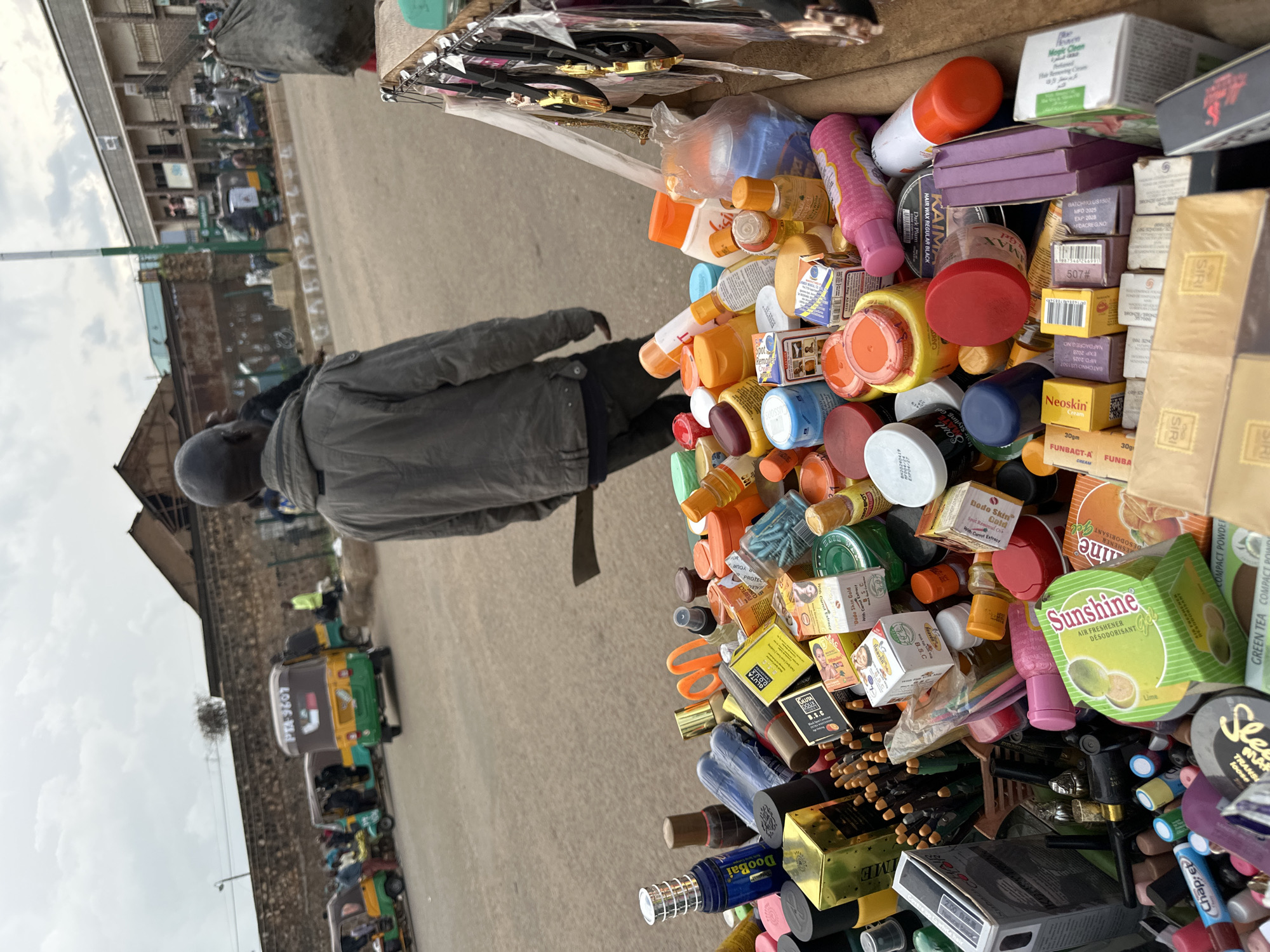 This is a close-up, eye-level outdoor shot of a vendor's stall overflowing with various cosmetic and personal care products. The stall is positioned on a paved street, with a blurry background of a busy street scene. In the background, there are several auto-rickshaws, known as "keke napep," some with passengers, and a few buildings. A man in a grey jacket is walking away from the camera, past the stall. The stall itself is a wooden cart filled with an assortment of items, including numerous small bottles, jars, tubes, and boxes of products. Some visible labels and packaging suggest items like perfumes, lotions, hair gels, compact powders, and air fresheners. The products are densely packed, with some items spilling over the edges of the cart. The overall impression is that of a vibrant, bustling market environment. The location is identified as Bukuru, Nigeria.