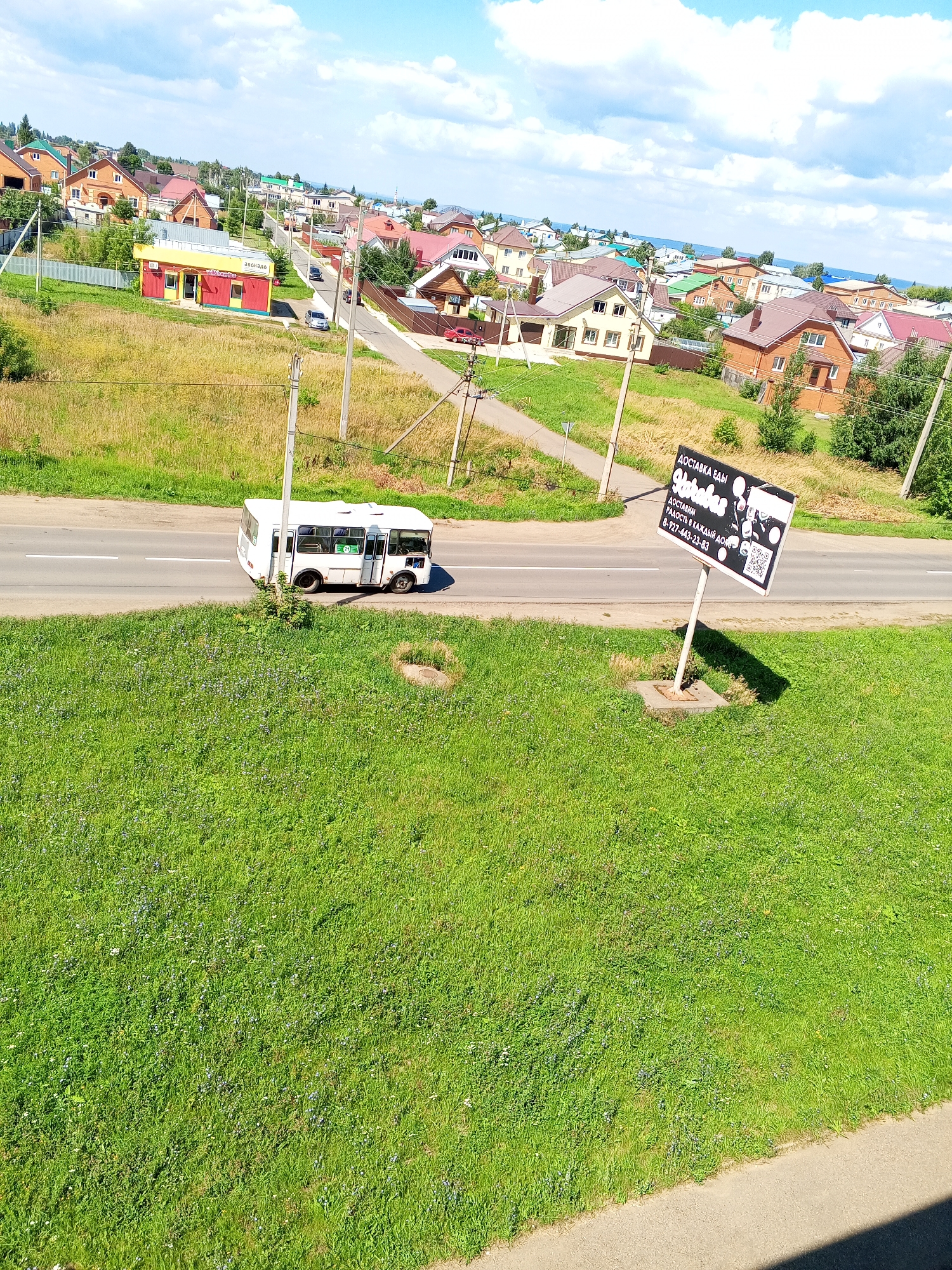 This is an elevated, medium-shot, full-color photograph of a suburban street scene. The image is dominated by a lush, green grassy foreground with scattered small wildflowers and a circular manhole cover in the lower right quadrant. A paved road runs horizontally across the middle of the image, with a white bus traveling on it from left to right. The bus is white and appears to be a minibus, with several windows and a door visible. To the right of the road, a large black sign with white text and graphics is mounted on a pole. The text on the sign appears to be in Russian and advertises food delivery, including a phone number and QR code. Behind the road, a collection of houses with various roof colors and architectural styles are visible on a gentle slope. Some of the houses are brick, while others are sided. A small, bright yellow and red building with a sign is located on the left side of the road. The background features more houses, trees, and a bright blue sky with scattered white clouds. The overall impression is of a sunny day in a residential area.