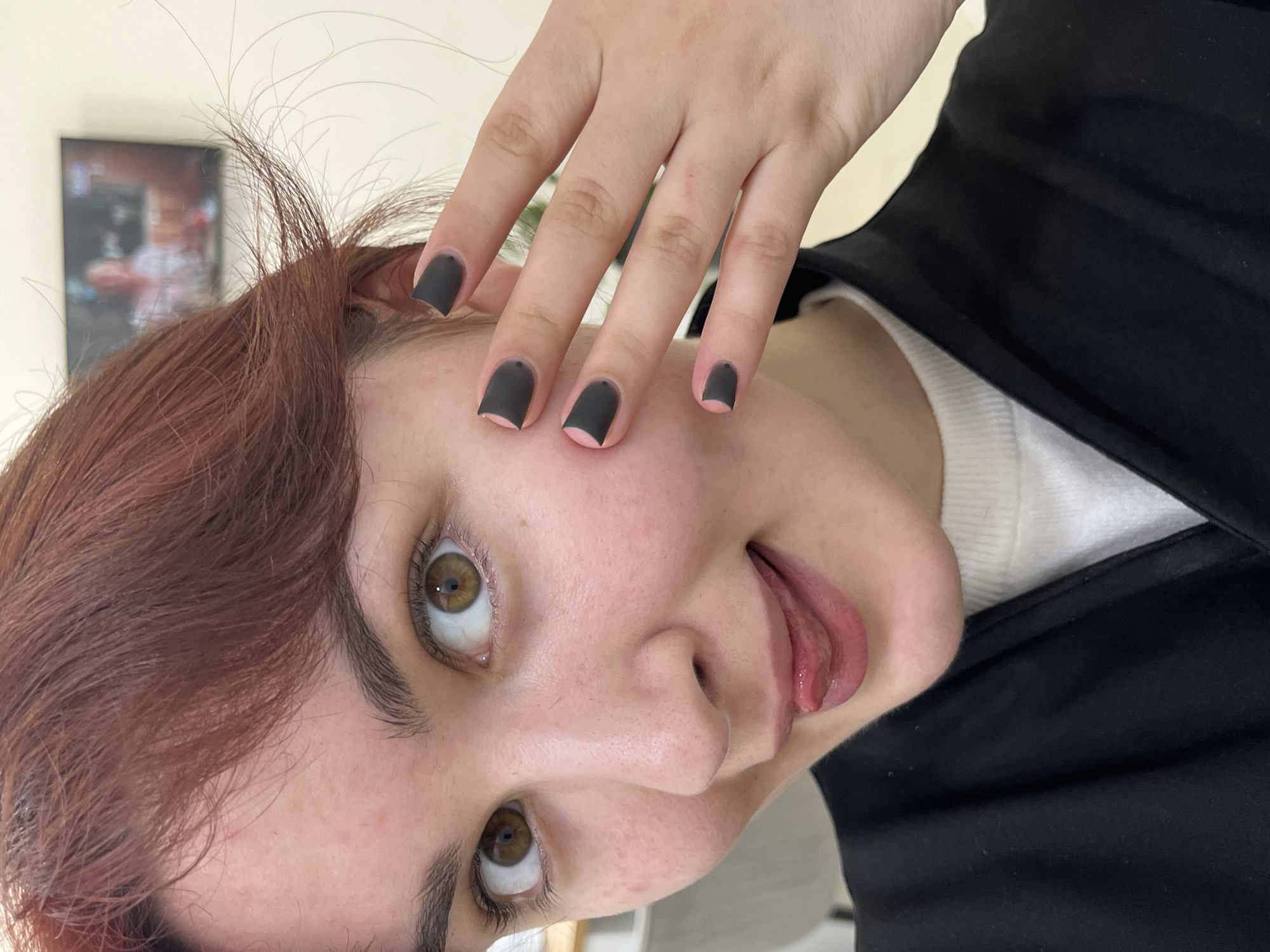 A person with reddish-brown hair and hazel eyes is positioned with their head tilted, sticking their tongue out playfully. Their hand is raised, displaying nails painted with a matte black color. They are wearing a white top and a black blazer. The background includes a picture on a wall and general interior features. There is no evidence in this image to indicate it was taken in Neftekamsk, Russia.