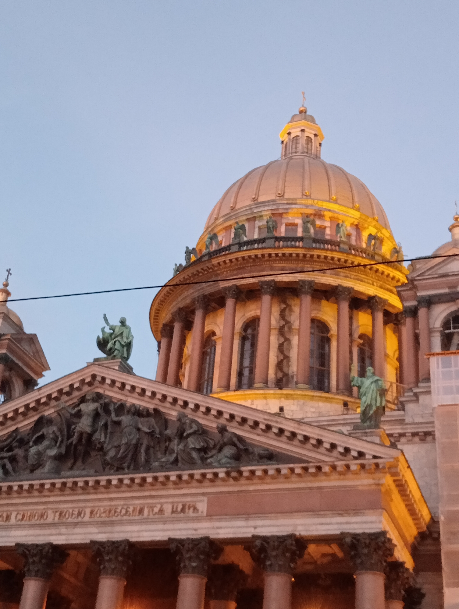 The image captures the dome and facade of Saint Isaac's Cathedral in Saint Petersburg, Russia, at dusk. The imposing golden dome is illuminated from below, casting a warm glow against the pale blue sky. The cathedral's architectural details, including its massive Corinthian columns, ornate friezes, and statues of figures in green patina, are visible. A line of text, likely an inscription, is present below a sculpted pediment. The overall scene conveys a sense of grandeur and historical significance.