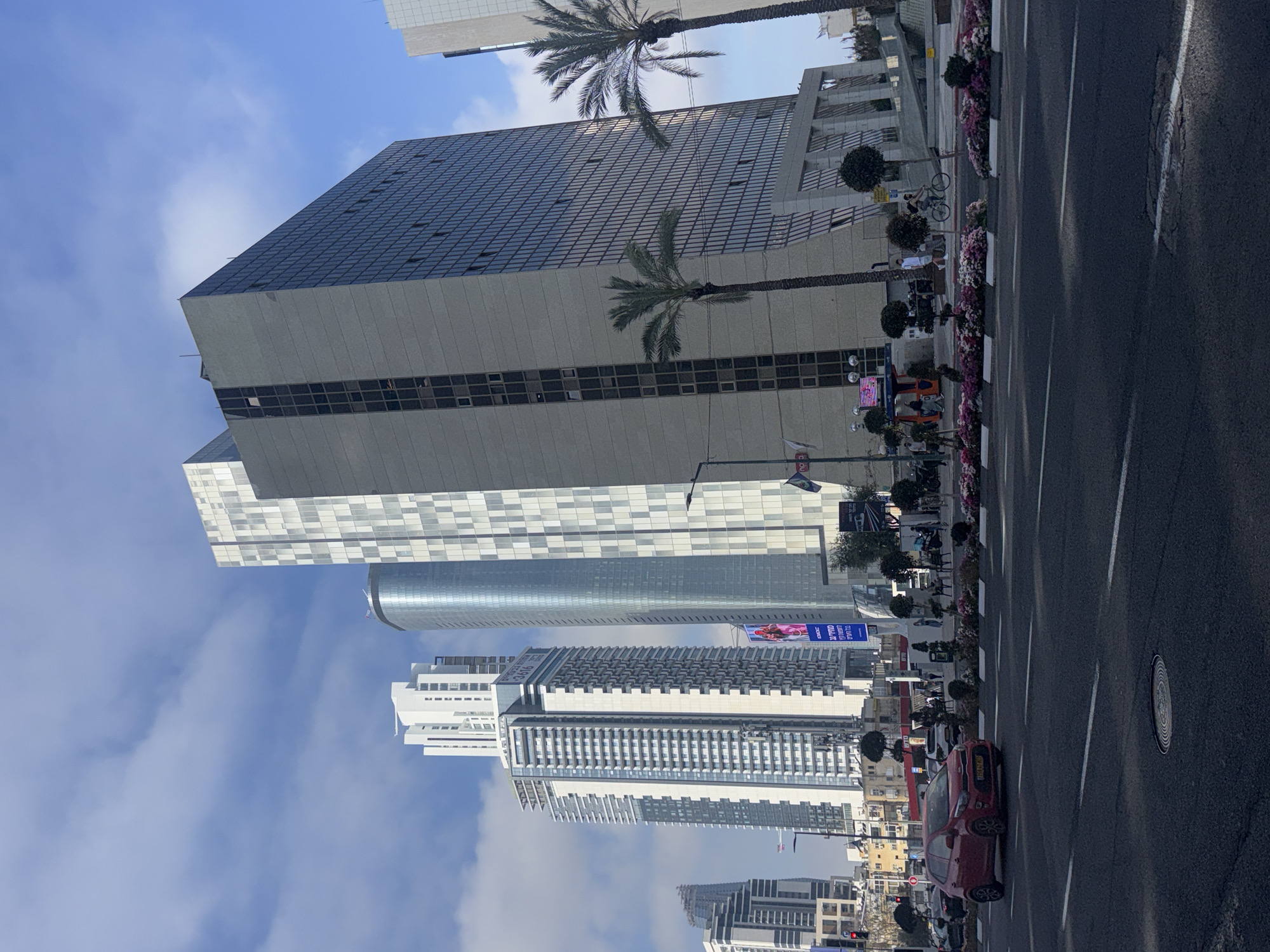 The image shows a street in Ramat Gan, Israel, lined with skyscrapers and palm trees. The street has a black asphalt surface and white lane markings. A red car is parked on the side of the road. Pedestrians are visible on the sidewalk near a row of flowering plants and small trees. The sky is blue with some clouds. The buildings are tall and modern, with varying facades.