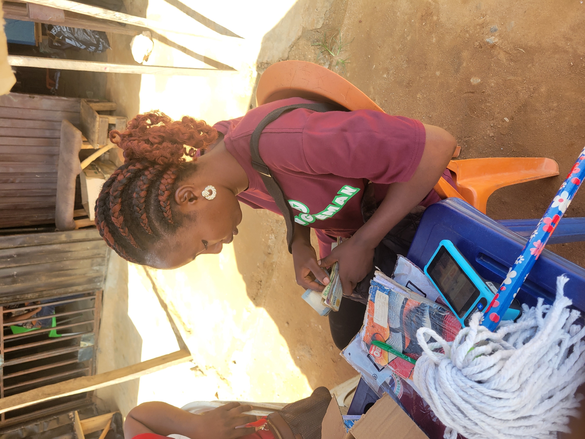 In Kwamba, Nigeria, a young woman with braided auburn hair is counting money, holding a wad of cash in her hands. She is wearing a maroon t-shirt with green lettering that appears to read "CHANGE MAN" and a sparkling earring in her left ear. She is seated on a blue plastic chair, with an orange chair visible behind her. On the blue chair, a mop with a floral-patterned handle, a stack of books or notebooks, and a small blue electronic device are placed. The scene appears to be outdoors, with a dirt ground and sunlight casting shadows, suggesting it is daytime. The background shows some wooden structures, possibly a market stall or dwelling, and a metal grate. The woman's expression is one of concentration as she handles the money.