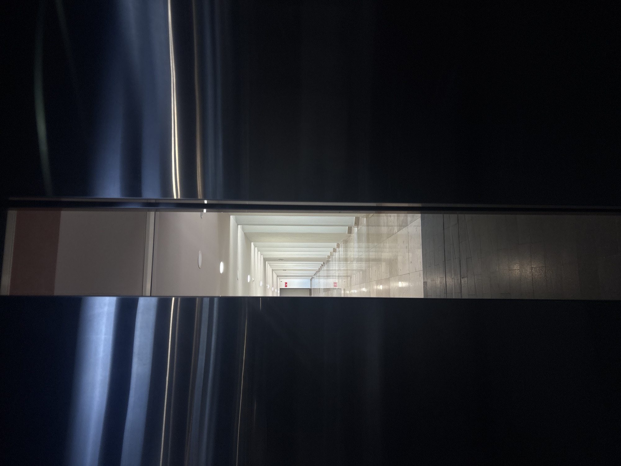 The image shows a view down a long hallway through a narrow opening. The hallway is brightly lit with a white ceiling and light-colored walls. There are evenly spaced lights along the wall. A red exit sign is visible in the distance. The floor appears to be tiled. The opening through which the hallway is viewed is framed by dark, reflective surfaces on the top and bottom.
The location of the image is not provided in the prompt and cannot be determined from the image itself.