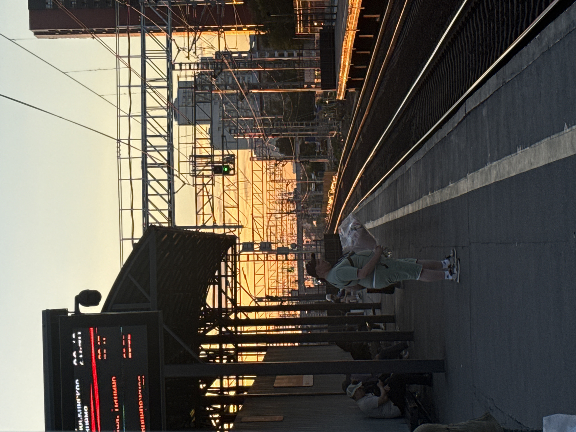 The image depicts a train station platform in Khimki, Russia, during a sunset. The warm, golden light of the setting sun illuminates the scene, creating a dramatic contrast with the shadows. In the foreground, a man is seen standing on the platform, holding a plastic bag over his shoulder. He is wearing a light green t-shirt and shorts, with sneakers on his feet. Another person is visible sitting further down the platform.
The platform itself is covered by a metal structure, and a digital display board showing train information is mounted on the left side of the image. The train tracks run alongside the platform, with the tracks themselves reflecting the sunset's glow. In the background, there is a complex network of overhead wires and scaffolding, suggesting ongoing construction or maintenance. A green traffic signal is visible on a mast among the overhead wires. The overall mood is calm and quiet, with the warm light evoking a sense of peacefulness. The visible text on the display board is primarily in Cyrillic characters.