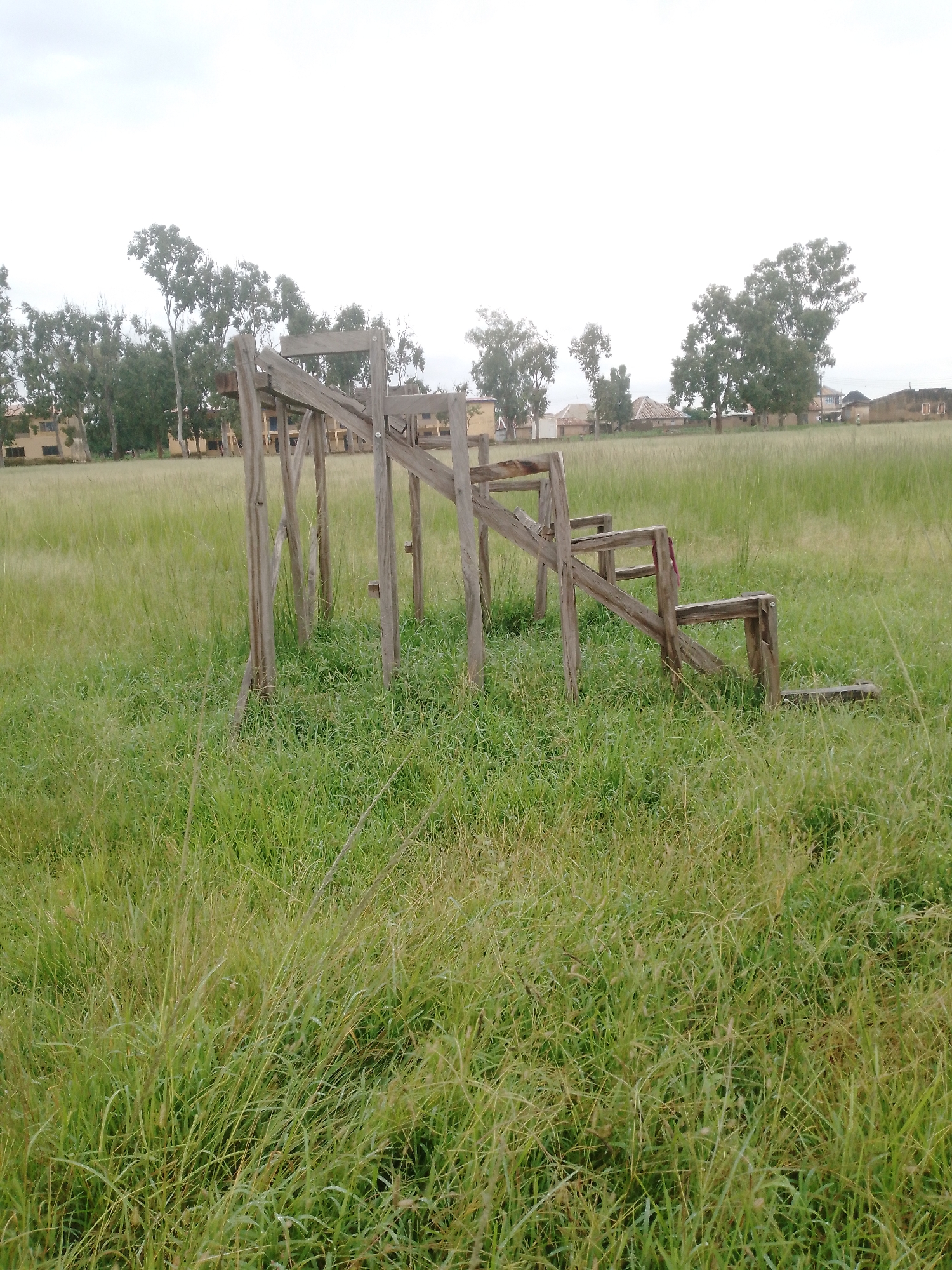 In the city of Jos, Nigeria, a weathered wooden structure resembling an unfinished playground climber or seating area stands in a field of tall, green grass. The structure, made of rough-hewn logs and planks, features a set of stairs leading up to a platform, with additional horizontal beams forming what could be supports or seating. The wood shows signs of age and exposure to the elements, with some pieces appearing cracked or splintered. In the background, a line of trees, including tall eucalyptus trees, separates the field from a cluster of buildings. The buildings appear to be low-rise, with light-colored facades and visible windows, suggesting a residential or institutional area. The sky above is overcast, casting a diffused light over the scene. The overall impression is one of a rural or semi-rural setting within the city, with elements of both nature and human construction present.