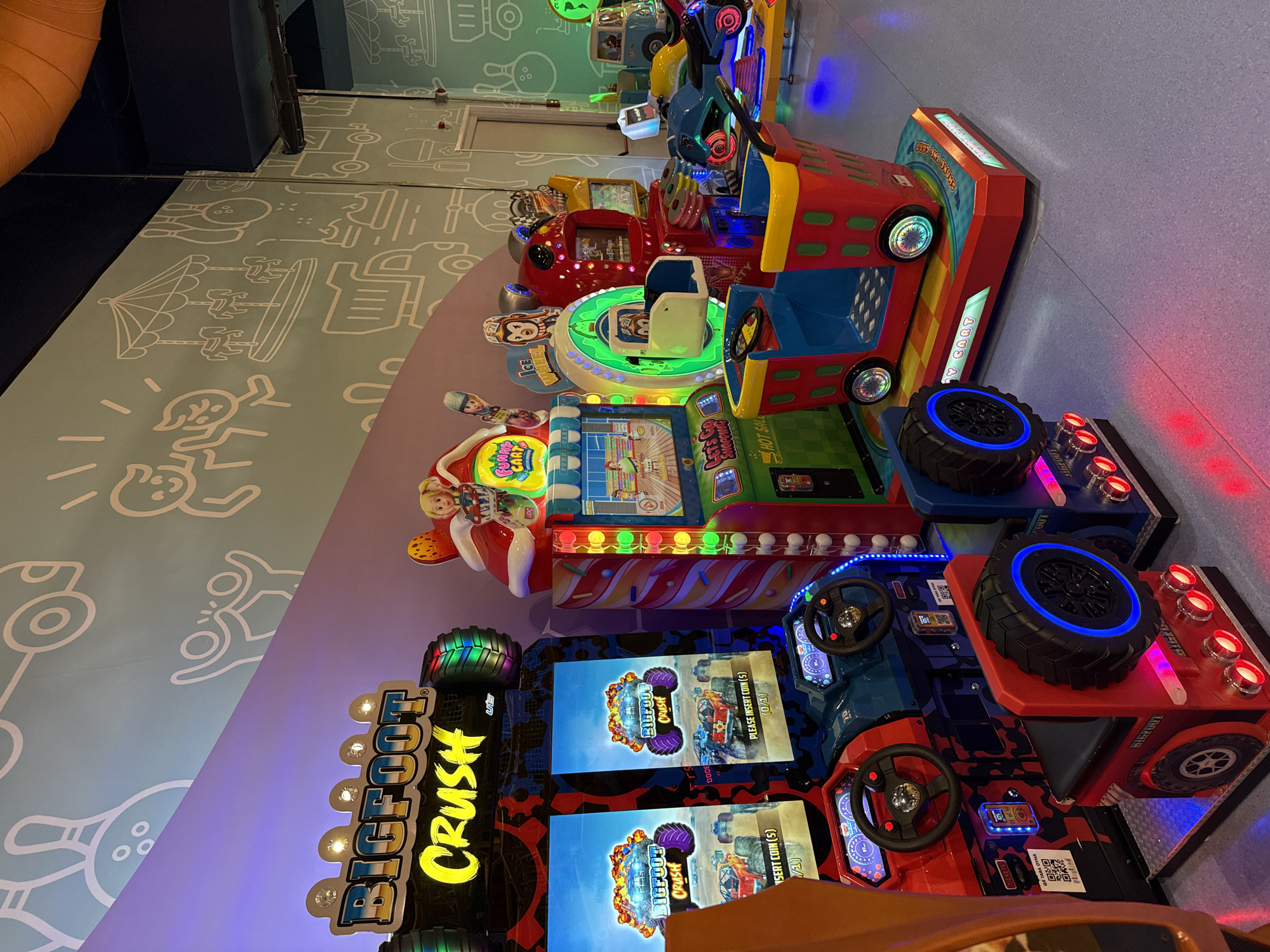 This is an indoor arcade with several brightly colored amusement machines. In the foreground, there are two "Bigfoot Crush" driving game machines with red and blue jeep-themed seats and steering wheels. Both machines have large screens displaying graphics of monster trucks. To the right of these, there is a "Funny Cart" ride with a red, yellow, and blue shopping cart-like seat. Further to the right, other arcade games are visible, including a motorcycle racing game and a game with a large green illuminated wheel. The walls are decorated with cartoonish line drawings of carnival rides, trucks, and people. The lighting is a mix of ambient arcade lights and the colorful glow from the machines. The floor is a light blue-gray. The scene appears to be in a family entertainment center or arcade, likely in a public space. The image does not provide any specific visual cues to confirm the location is Üsküdar, Turkey.