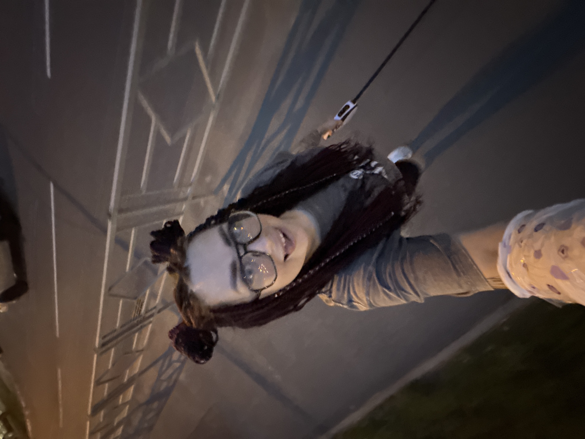 A first-person perspective shot captures a woman with purple-streaked braids styled into two buns, wearing large glasses and a grey t-shirt, as she holds a leash for a dog, indicated by the leash extending out of frame and a light on the handle. The woman is standing on asphalt, with white lines marking a roadway visible in the background, suggesting an urban environment. A metal fence runs parallel to the roadway, casting long shadows on the ground. The scene appears to be in the city of Tolyatti, Russia, likely a street or park at dusk or night, given the artificial lighting and shadows. The woman's expression is joyful, with her mouth open and tongue slightly out.