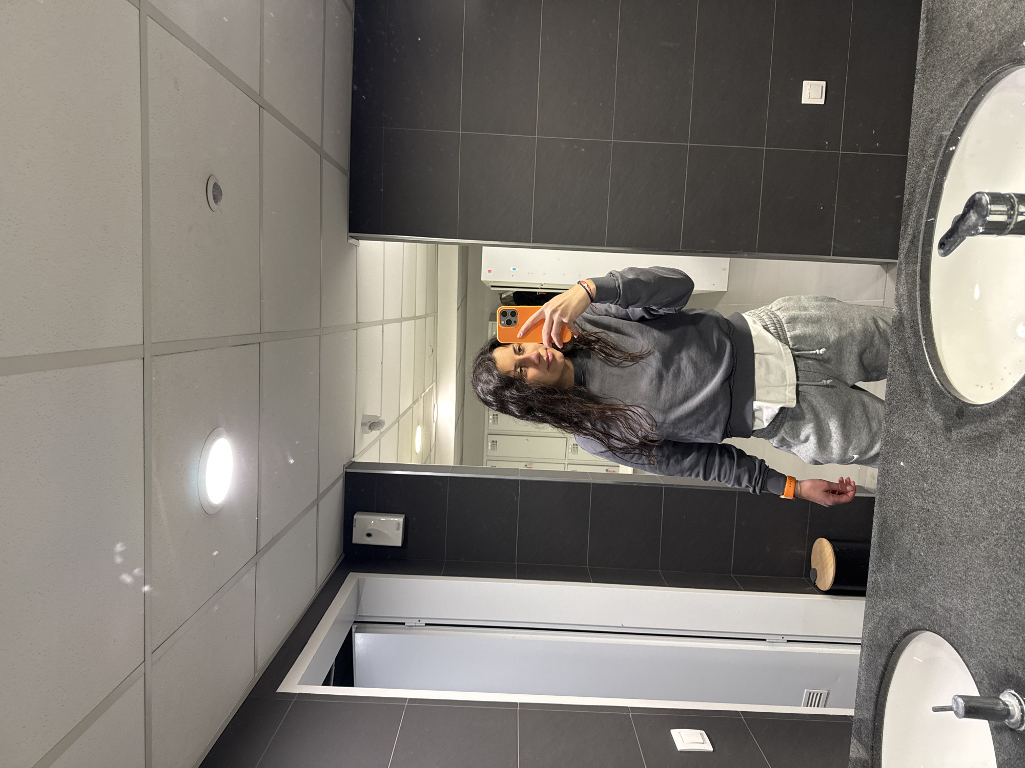 A young woman is taking a selfie in a public restroom. The restroom has white tiled ceilings with recessed lighting, and dark grey tiled walls. There are sinks visible in the foreground. In the background, a mirror reflects the woman holding a phone and rows of lockers. The exact location within the city of Unknown, Unknown, is indistinguishable.