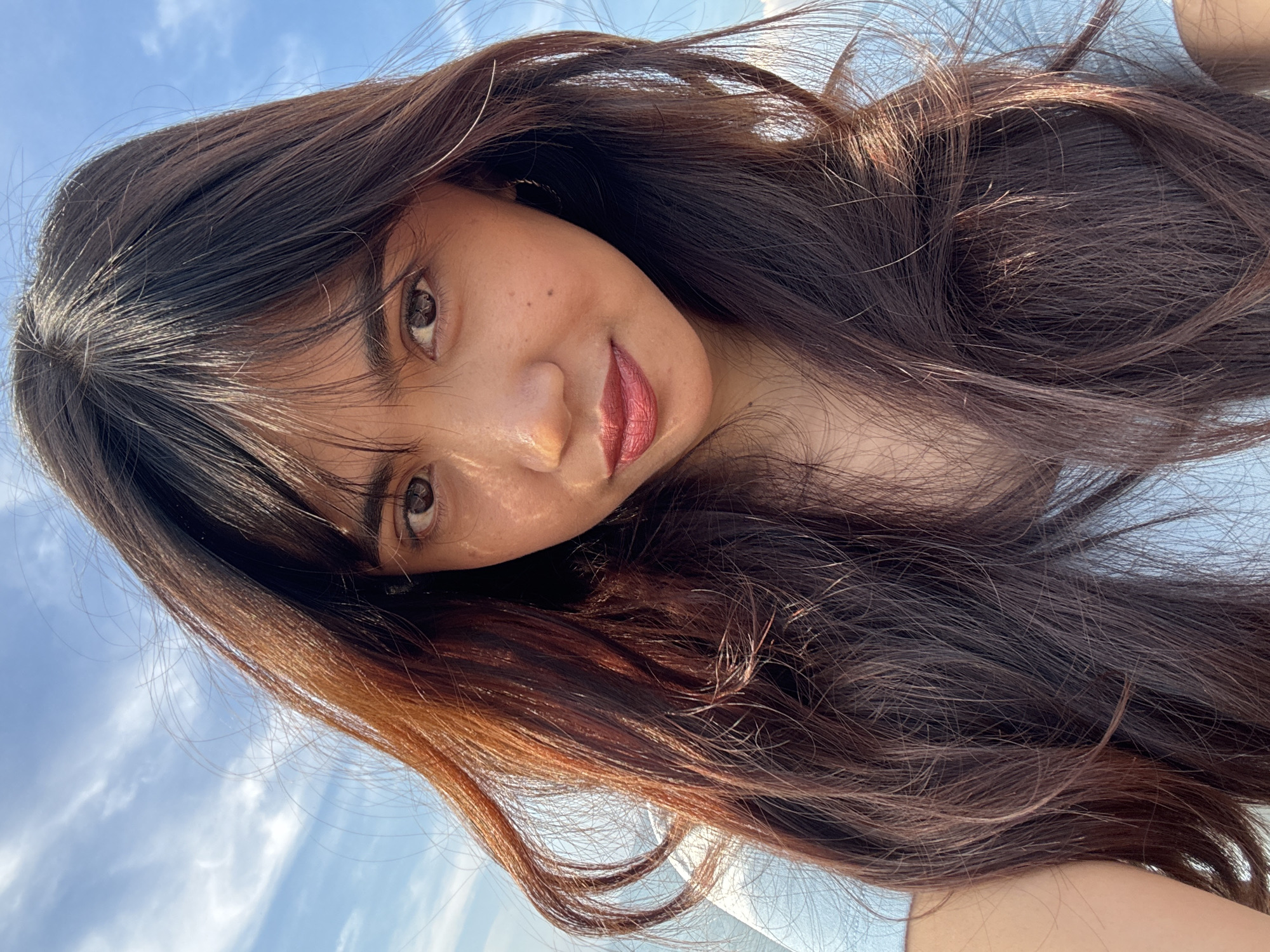 A young woman with long, dark brown hair is shown in a close-up selfie against a bright blue sky with wispy clouds. Her hair is blowing slightly in the wind. She has fair skin with a few freckles and is wearing dark lipstick. Her eyes are brown and she is looking directly at the camera. The background suggests an outdoor setting, possibly a beach or open area, consistent with locations in Davao City, Philippines.