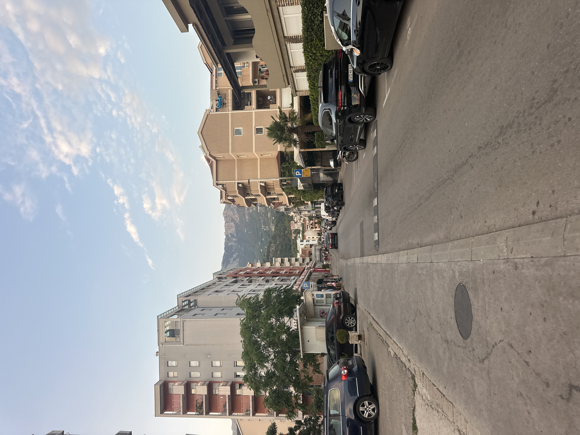 The image shows a street in Budva, Montenegro, with multi-story buildings lining both sides of the road. The buildings are a mix of residential and commercial structures, with balconies and windows visible on their facades. Several cars are parked along the street, including SUVs and sedans. People can be seen walking on the sidewalk, and a few are seated at outdoor tables. In the background, mountains rise above the city, and the sky is partly cloudy. The scene appears to be captured during the late afternoon or early evening, with the sun casting a warm glow on the buildings. The street is paved with asphalt, and a sidewalk runs along the left side of the road. A speed bump is visible on the right side of the street.
