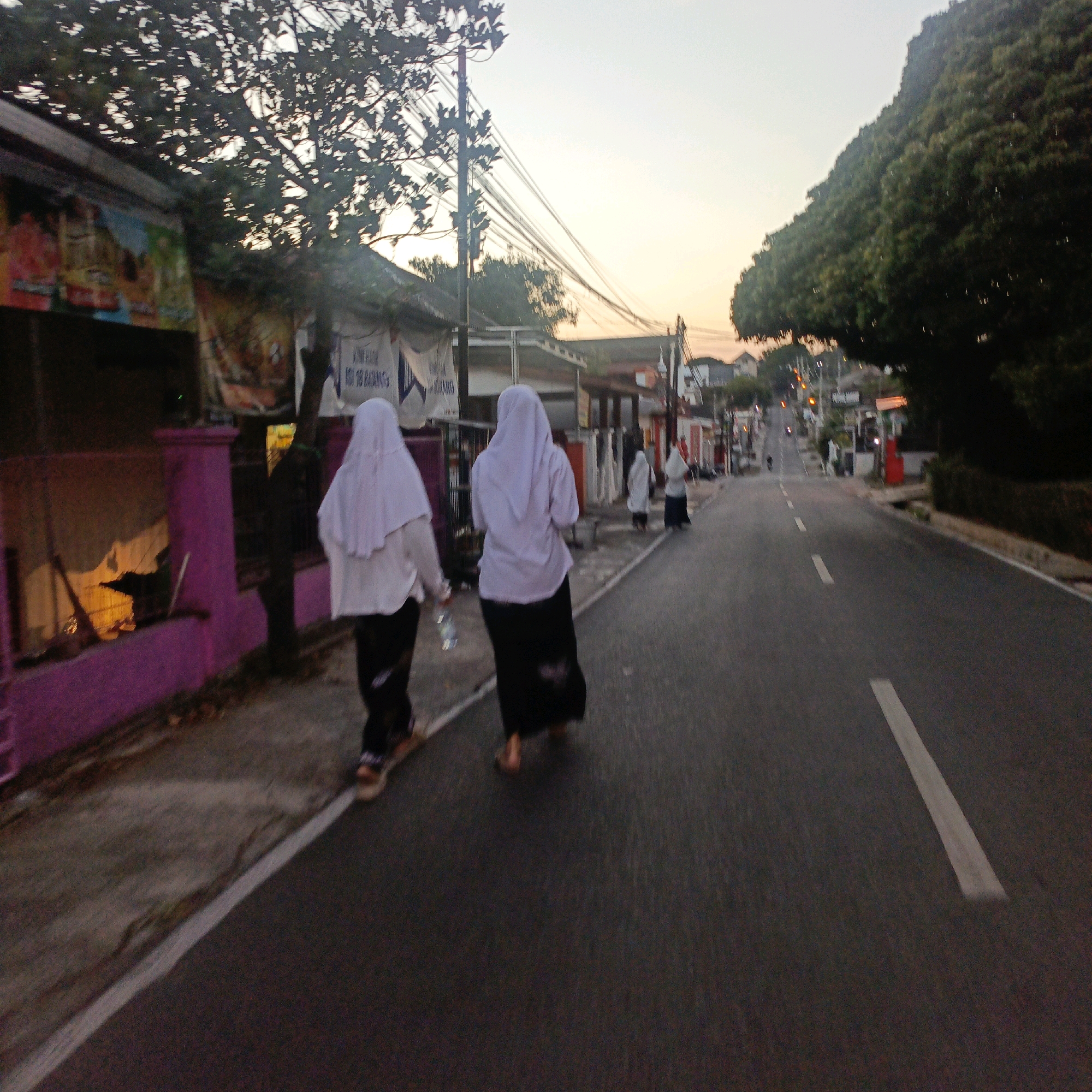 The image depicts a street scene in Salatiga, Indonesia, at dusk. Two individuals, seen from behind and walking away from the viewer, are the primary subjects. They are both wearing white hijabs and long dark skirts, suggesting they are Muslim women. The person on the left is holding a clear plastic bottle. They are walking on the sidewalk adjacent to a road.
To the left of the sidewalk, there is a low wall painted bright pink, with some indistinct signage or posters on a building behind it. A tree with dense foliage is visible on the left side of the frame, partially obscuring the sky and the buildings. Overhead, a network of electrical wires stretches across the scene, supported by poles.
The road itself is asphalt and has a white line marking its edge. Further down the road, the scene shows other buildings and trees, as well as more people walking in the distance. The sky is a pale orange and yellow, indicative of sunset. The overall lighting suggests it is either early morning or late afternoon. The environment appears to be a mix of residential and commercial buildings, with a distinctly Indonesian character.