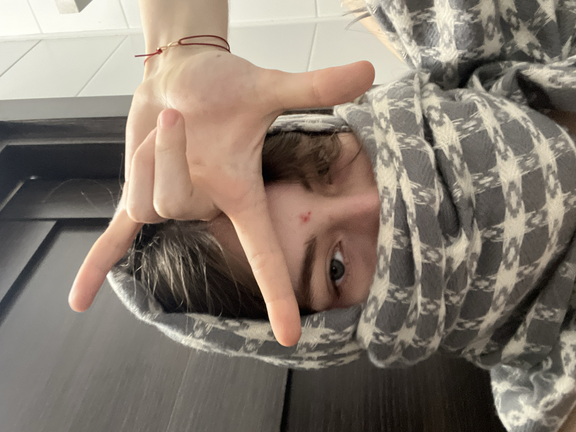 The image captures a close-up, inverted view of a person indoors. The individual's head, angled downwards, is partially enveloped by a grey and white patterned scarf or blanket, which features a distinct houndstooth-like design interspersed with abstract star shapes. Only the right eye, eyebrow, and a portion of the forehead and nose bridge are visible, revealing a calm gaze directed towards the camera. A small red blemish is noticeable on the forehead.
The person's left hand is prominent in the foreground, raised towards the camera. The fingers are arranged with the thumb and pinky extended, while the index and middle fingers are bent, creating a distinct 'L' shape or a framing gesture around the face. A thin red string bracelet with a small metallic bead adorns the person's left wrist.
The background consists of indistinct indoor elements: a dark, flat surface occupies the lower and left portions of the frame, possibly a door or wall panel, contrasting with a lighter, possibly paneled ceiling or upper wall section in the top left. The photograph presents a casual pose with an intimate and slightly disorienting perspective, consistent with an indoor setting in Saint Petersburg, Russia. No visible text is present.