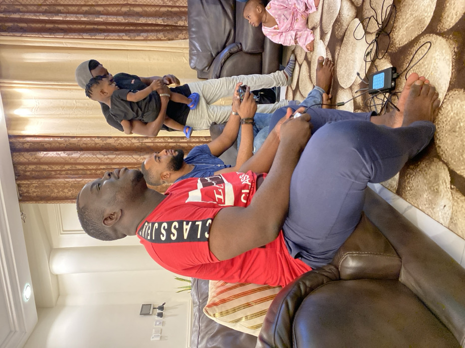 The image, rotated 90 degrees clockwise, captures a casual indoor scene, likely a living room, in Adenta Municipality, Ghana. Several individuals, primarily males, are present.
In the foreground, a man wearing a red t-shirt with "CLASS JEANS" printed on it is seated on a brown sofa, engaged in playing a video game with a controller in his hands. Beside him, another man in a blue t-shirt is also seated and holding a gaming controller, looking towards the right, presumably at a screen beyond the frame. Both men are barefoot.
In the mid-ground, a man wearing a grey hoodie or cap is holding a young child dressed in a black top. They appear to be observing the gaming activity. On the patterned carpet to the right, a baby or toddler in a pink patterned sleepsuit is sitting, also looking towards the right.
On the floor, between the gamers and the baby, a small electronic device, likely a gaming console or mini-PC, is visible with multiple connecting wires. The room is furnished with brown leather-like sofas and features patterned gold and brown curtains in the background. Indoor lighting fixtures are visible on the ceiling. The scene depicts a relaxed gathering, possibly a family enjoying leisure time together with video games.