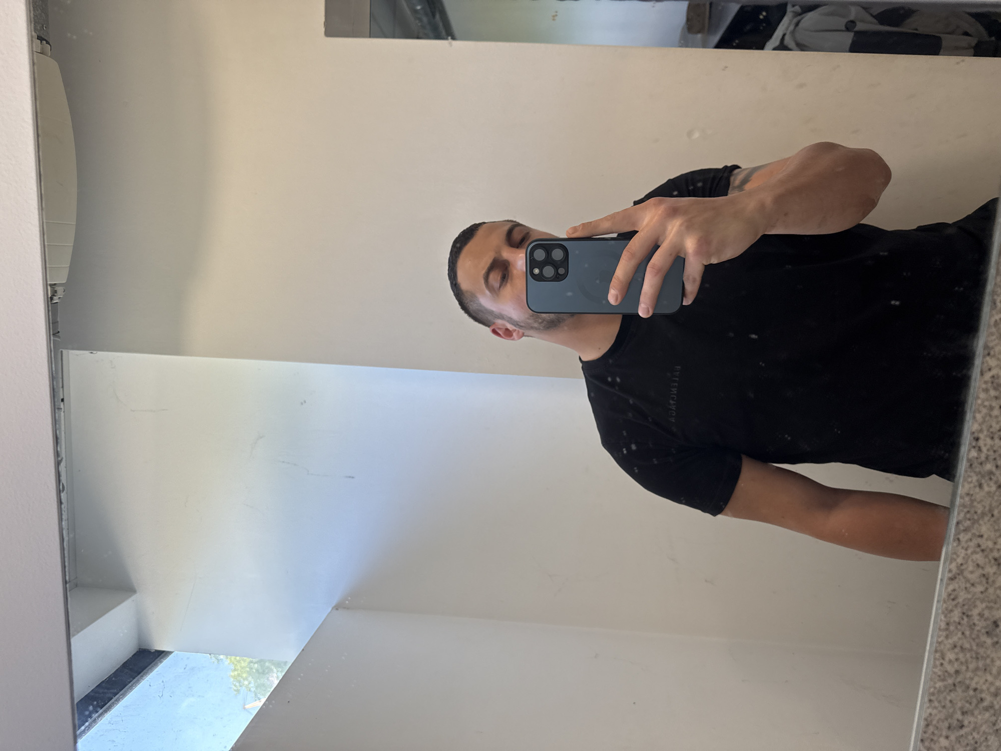 A person is taking a selfie in a mirror. The person is a man with short hair and is wearing a black shirt. He is holding a blue smartphone. The background appears to be a bathroom with white walls. There is no information in the image to indicate this location is in Varna, Bulgaria.